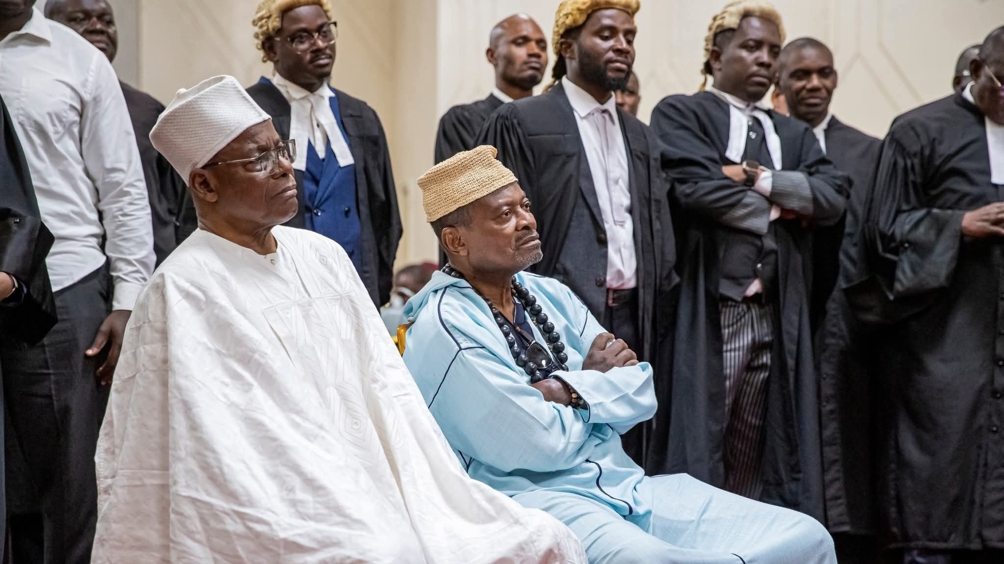 The image shows a group of individuals in formal attire, likely in a courtroom or legal setting. Two prominent figures in the foreground are seated, dressed in traditional attire. The person on the left wears a white outfit and a hat, while the individual on the right is in an aqua-colored garment with a distinctive headpiece. Behind them, several people are standing, some in legal robes, suggesting a gathering of lawyers or officials. The atmosphere appears serious and formal, indicating an important event or proceedings.