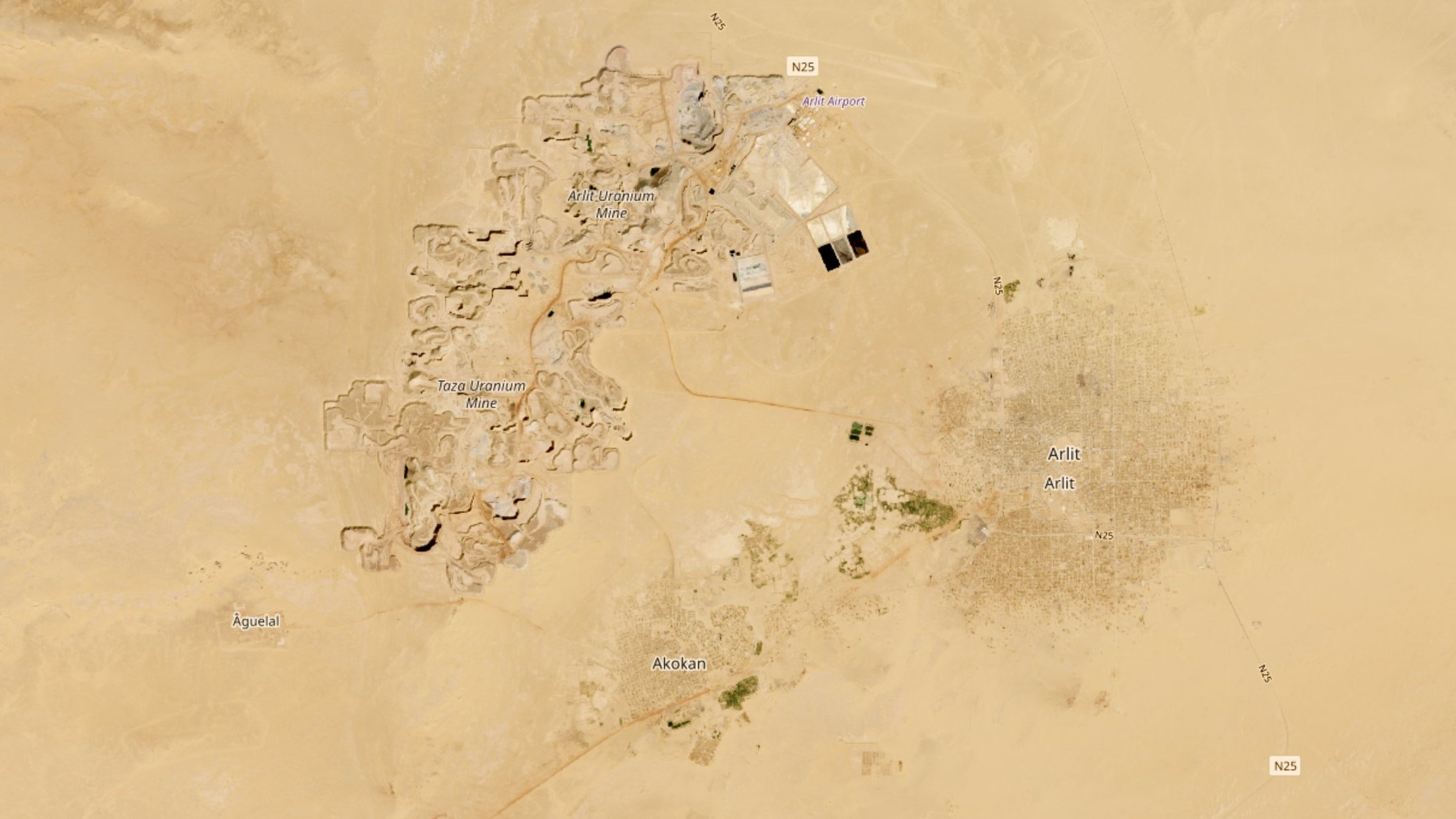The image appears to be an aerial view of a desert landscape featuring two distinct areas. On the left side, there are ruins or archaeological sites, indicated by clusters of structures, possibly remnants of ancient buildings. Nearby, there is an airport and some roads connecting the areas. On the right side, there is a more densely populated area, showing what looks like a town or settlement named "Antic," characterized by a grid-like pattern, suggesting regular streets and organized layout. Surrounding the settlement, there are patches of greenery, likely indicating agricultural activity. The vast surrounding landscape is primarily sandy and barren, typical of desert regions.