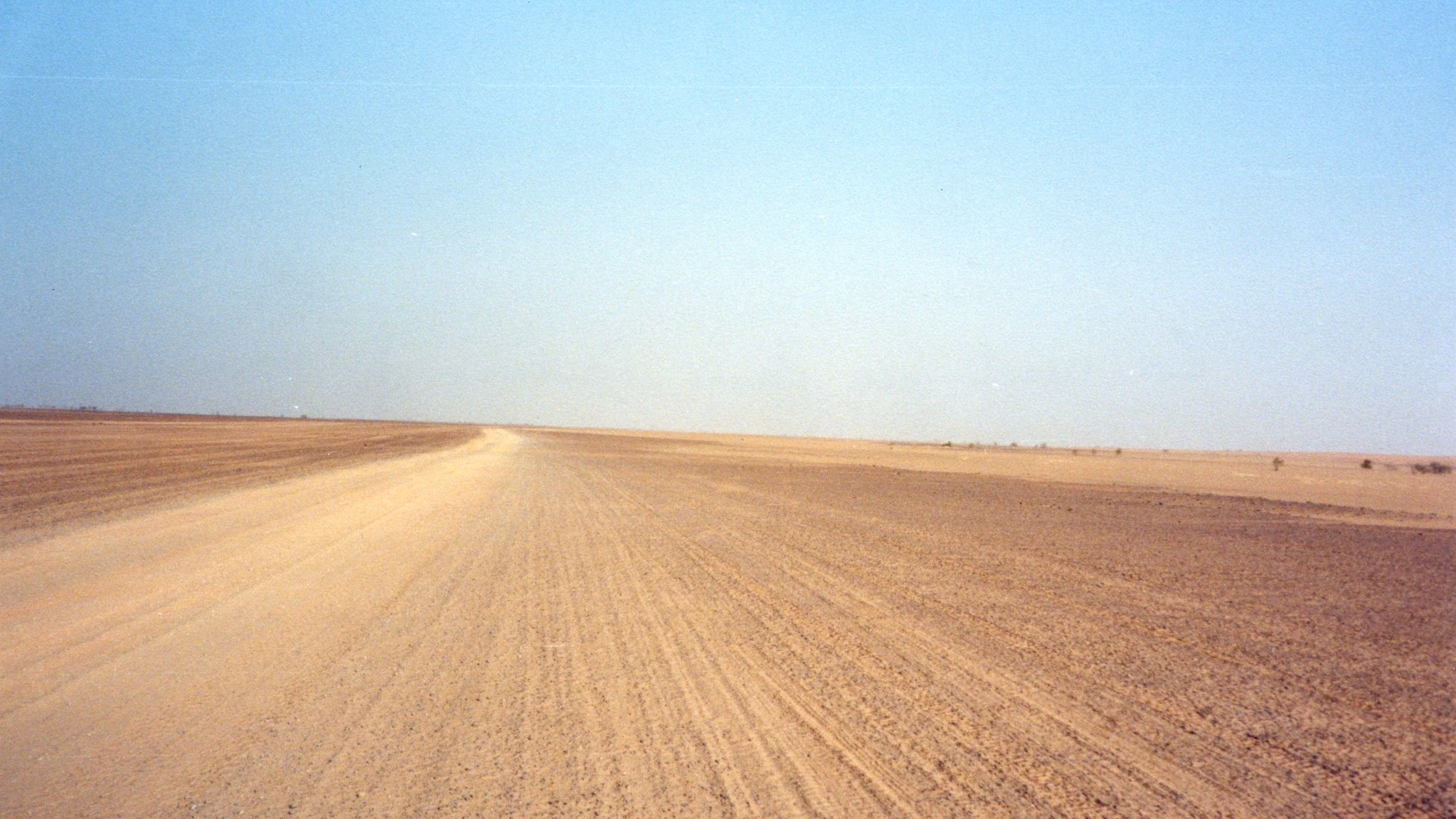 The image depicts a barren landscape characterized by a vast, empty stretch of land, likely a desert or arid region. The foreground features a dirt road that leads into the distance, flanked by dry, flat terrain. The sky overhead is clear and blue, creating a stark contrast with the earthy tones of the ground. The overall scene conveys a sense of isolation and openness, typical of remote, dry environments.