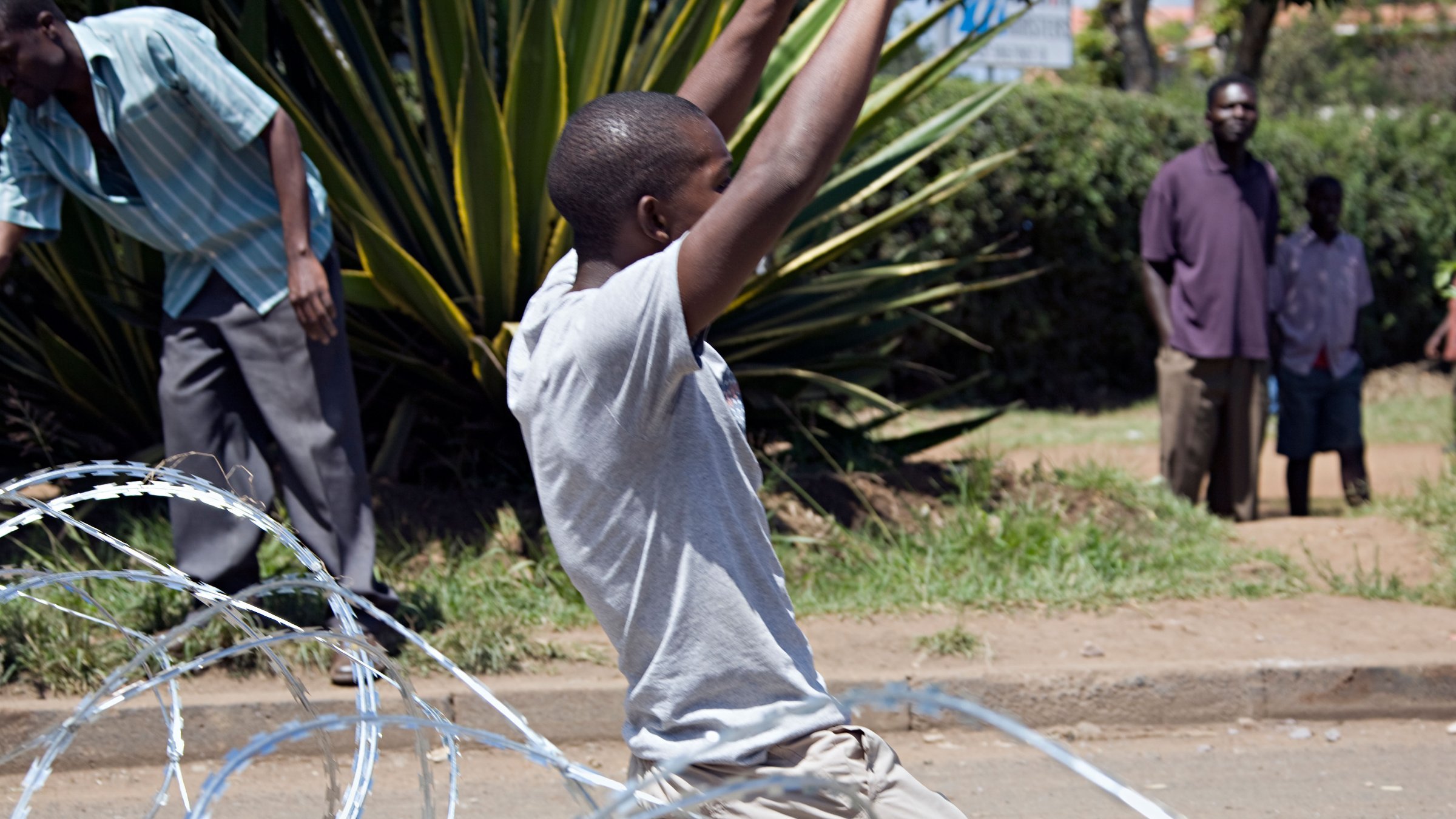 The image depicts a scene where a young man is on his knees, appearing to be in a moment of distress or surrender. He is surrounded by coils of barbed wire on the ground. In the background, there are several individuals observing the situation, some appearing to be standing casually while others are more focused on what is happening. The setting looks to be an outdoor area, possibly a street, with greenery in the background. The overall atmosphere seems tense and conveys a sense of conflict or protest.