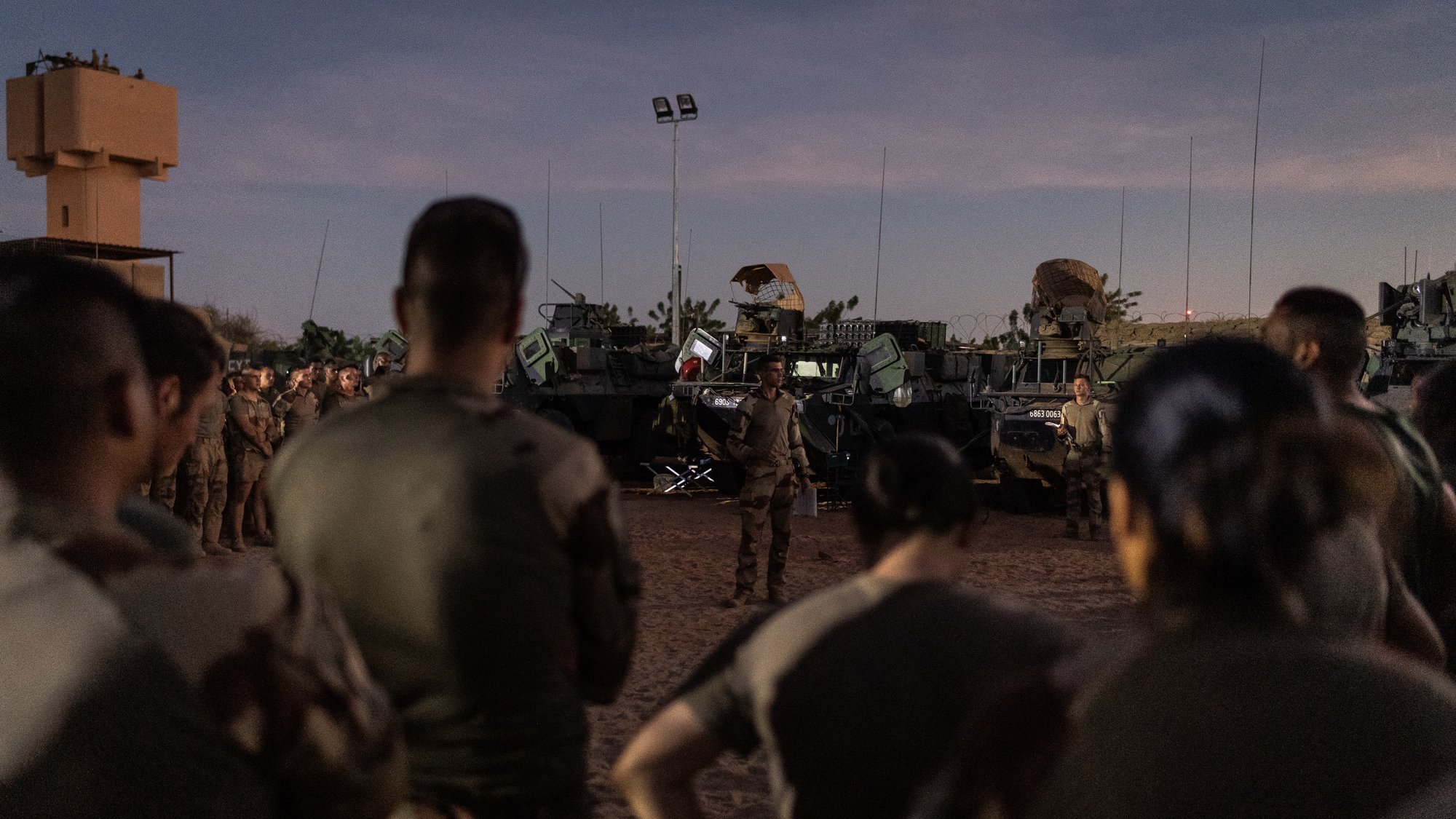The image depicts a group of military personnel gathered in a semi-dark environment, likely during dusk. They are standing in front of several military vehicles, which appear to be armored and equipped for operational purposes. A leader, possibly giving a briefing or instructions, is positioned in front of the group. The scene conveys a sense of preparation or briefing before a mission or operation. The setting indicates a desert-like terrain, typical for military exercises or deployments in such environments.