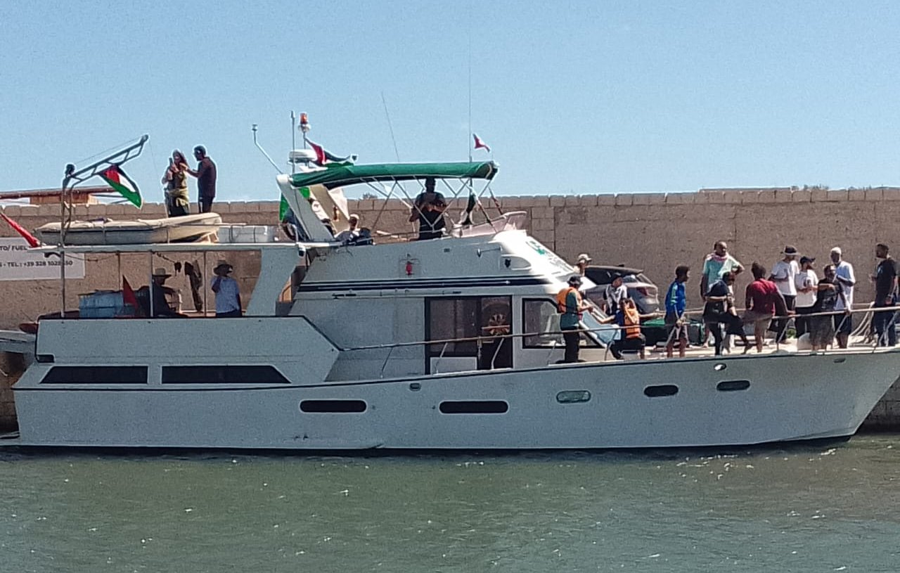 The image shows a large motorboat docked at a marina. The boat has a white hull with a green and white canopy on top. A group of people can be seen on the boat and along the dock, some engaging in conversation and others preparing to board. The surrounding area appears sunny, and there are flags displayed on the boat and nearby structures. The water is calm, reflecting the bright blue sky.