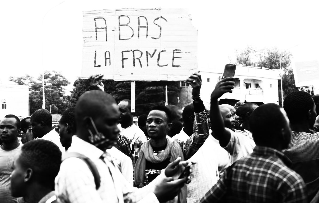 L'image représente une scène de manifestation. Au premier plan, plusieurs personnes se rassemblent, certaines levant les bras. Un homme, au centre, tient une grande pancarte avec l'inscription "ABAS LA FRYCE" en lettres capitales. La foule semble engagée et énergique, exprimant une certaine frustration ou un message de contestation. L'ambiance est chargée d'émotion, avec des visages déterminés et des gestes expressifs, dans un cadre urbain qui suggère un lieu public. Les vêtements des manifestants varient, ajoutant à la diversité de la scène.