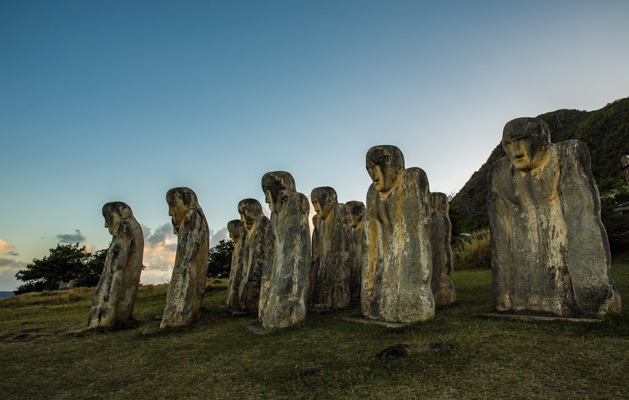 The image features a series of large stone sculptures arranged in a row, resembling human figures. These sculptures have distinct, elongated shapes and facial features, with some appearing to look downward. The background includes a grassy area and a hillside, with trees visible nearby. The sky above is clear, transitioning from blue to lighter tones as it approaches the horizon, suggesting either dawn or dusk. The overall atmosphere gives a sense of mystery and historical significance to the figures.