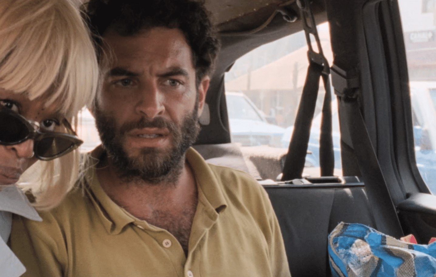 The image shows two people inside a vehicle. One person, likely wearing a blonde wig and sunglasses, leans towards the other, who has a beard and a concerned expression. The setting appears casual and slightly cramped, with a background that suggests they are parked in a busy area. The overall mood seems tense or serious, as both individuals have focused looks on their faces.
