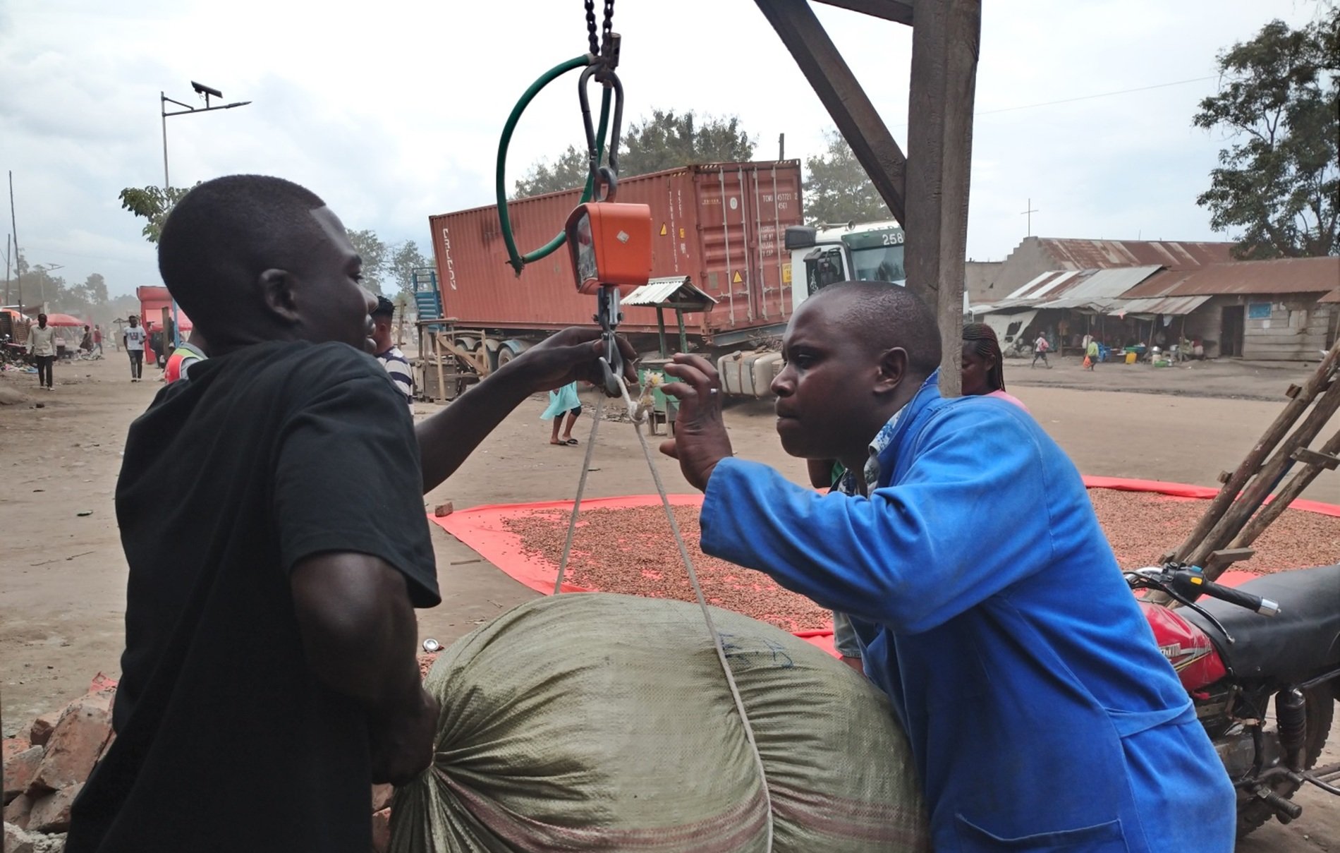 The image depicts a scene where two men are engaged in an activity involving a large sack. One man, wearing a blue coat, appears to be operating a scale or a weighing device to measure the weight of the sack being lifted by another man in a black shirt. The background shows a street with several structures and vehicles, suggesting a marketplace or an industrial area. There are also other people and containers visible in the background, contributing to the bustling atmosphere of the scene.