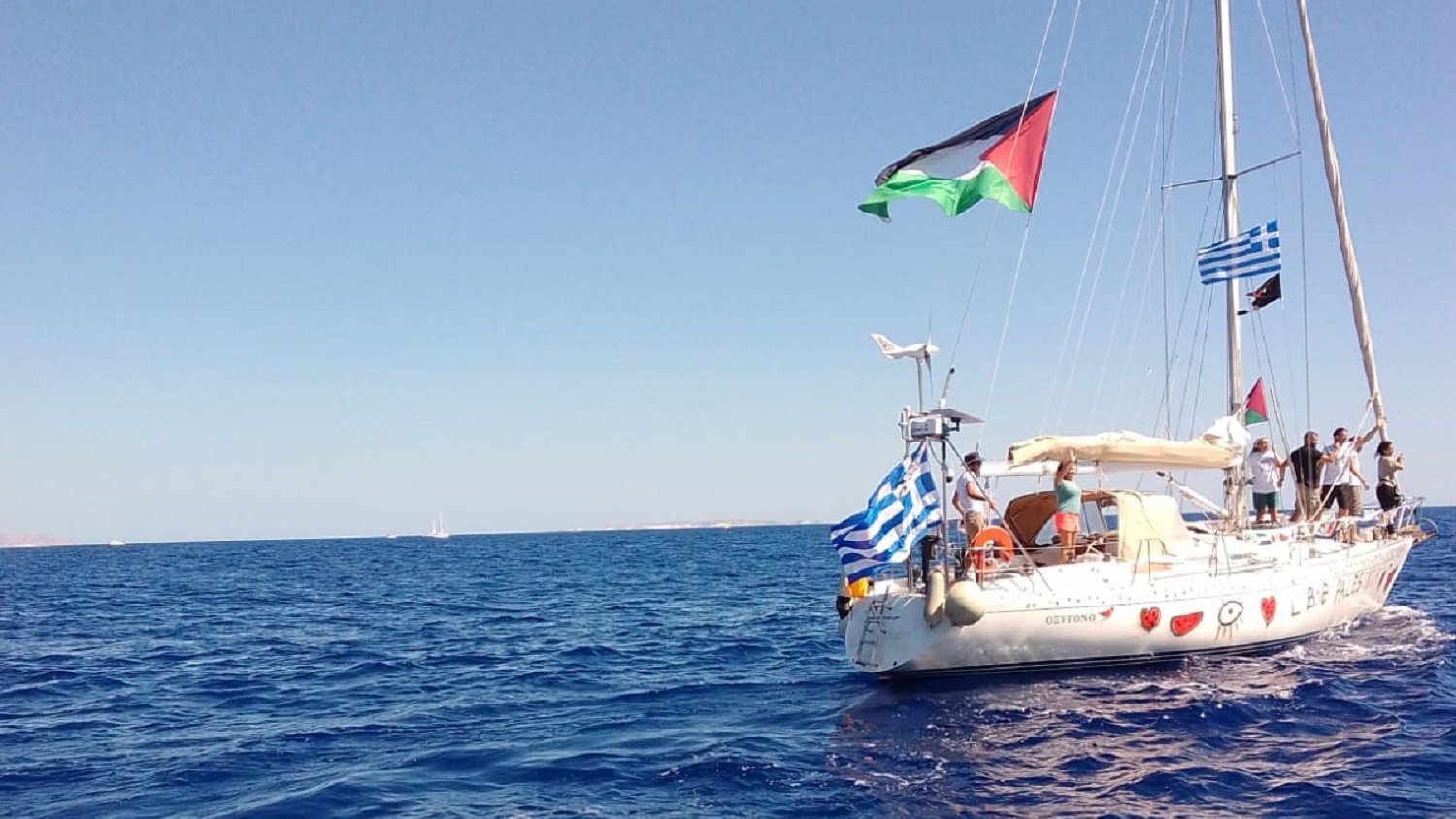 The image depicts a sailboat on calm blue waters. The boat features several flags flying from its mast: notably, a Palestinian flag, a Greek flag, and a blue-and-white striped flag typical of Greece. The sky is clear and bright, suggesting a sunny day at sea. The surrounding water appears serene, reflecting the sunlight.