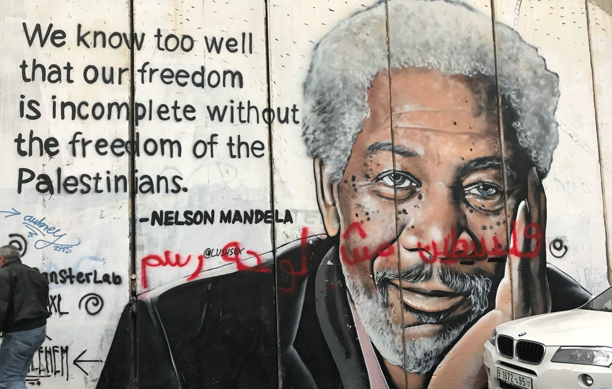 The image depicts a mural on a concrete wall, featuring a portrait of a man with gray hair and a thoughtful expression. The mural also includes a quote attributed to Nelson Mandela that reads, "We know too well that our freedom is incomplete without the freedom of the Palestinians." The background is a textured concrete surface, and there are some graffiti elements in red. The scene suggests themes of solidarity and freedom, particularly regarding the Palestinian struggle.