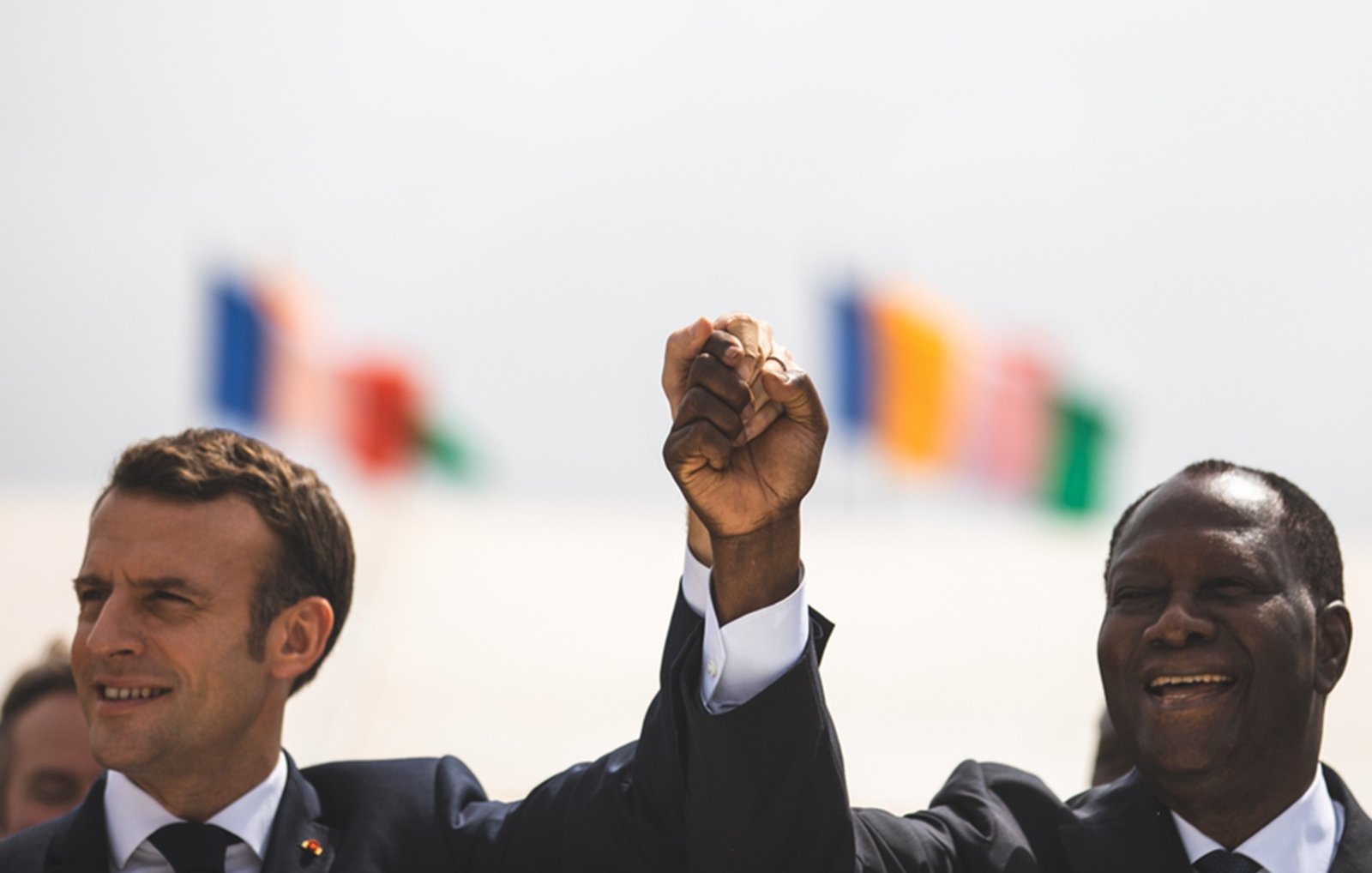 The image shows two men, one wearing a suit with a tie, raising their hands together in a gesture of solidarity or celebration. They are smiling and appear to be in a public setting. In the background, there are several flags visible, suggesting an international context or event. The atmosphere seems positive and hopeful.