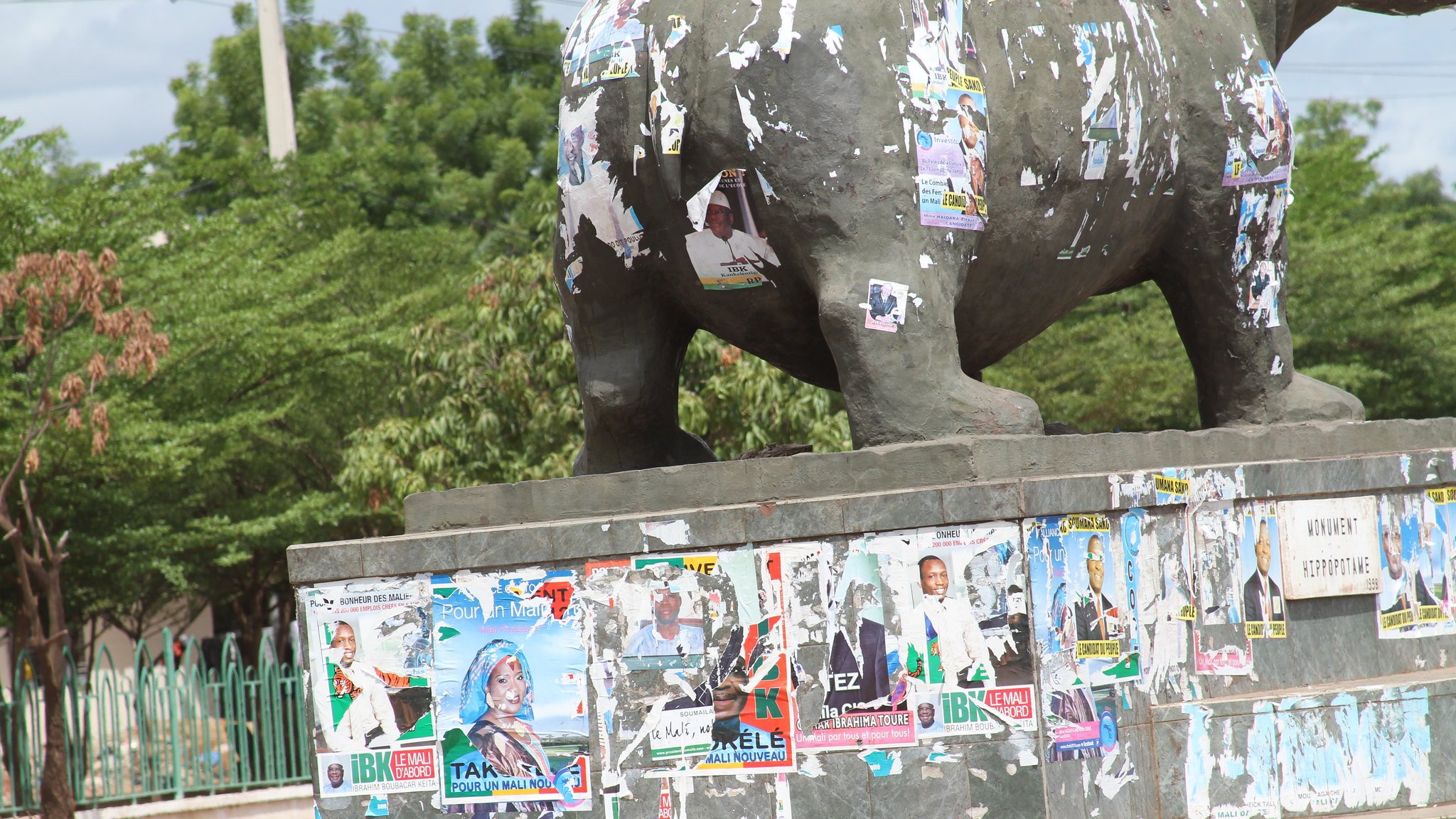 The image depicts a statue, likely an elephant, that is visibly weathered and covered in numerous political posters and flyers. These posters, in various colors, are peeling and overlapping, suggesting a history of political campaigns or events. The statue is situated in an outdoor area, surrounded by greenery and a fence in the background. The overall scene conveys a sense of urban life and the passage of time through the accumulation of posters.