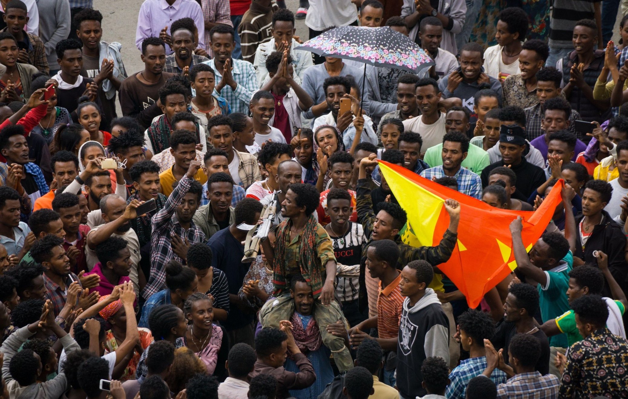 The image depicts a large gathering of people in a vibrant setting, likely representing a celebration or demonstration. Crowds are tightly packed together, with some individuals holding colorful flags, possibly indicating a cultural or political event. One person stands out in the center, engaging the crowd, which looks enthusiastic and expressive. The mood appears lively, with a sense of unity among the participants. Various expressions and clothing styles suggest a diverse group, reflecting a shared purpose or message. An umbrella can be seen, hinting at the weather conditions. Overall, it captures a moment of collective energy and communal spirit.