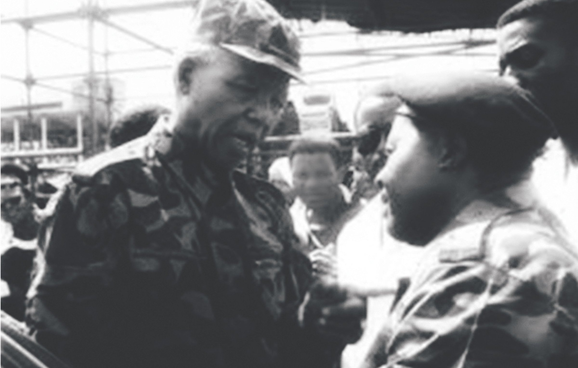 The image appears to be a historic black-and-white photograph depicting two men engaged in a conversation. One man is wearing military camouflage and appears to be in a position of authority, while the other is also dressed in military attire. They are surrounded by a group of people, indicating a significant setting, possibly a public event or gathering. The expressions on their faces suggest a serious discussion. The context hints at a moment of political or military importance, likely related to South Africa's history.
