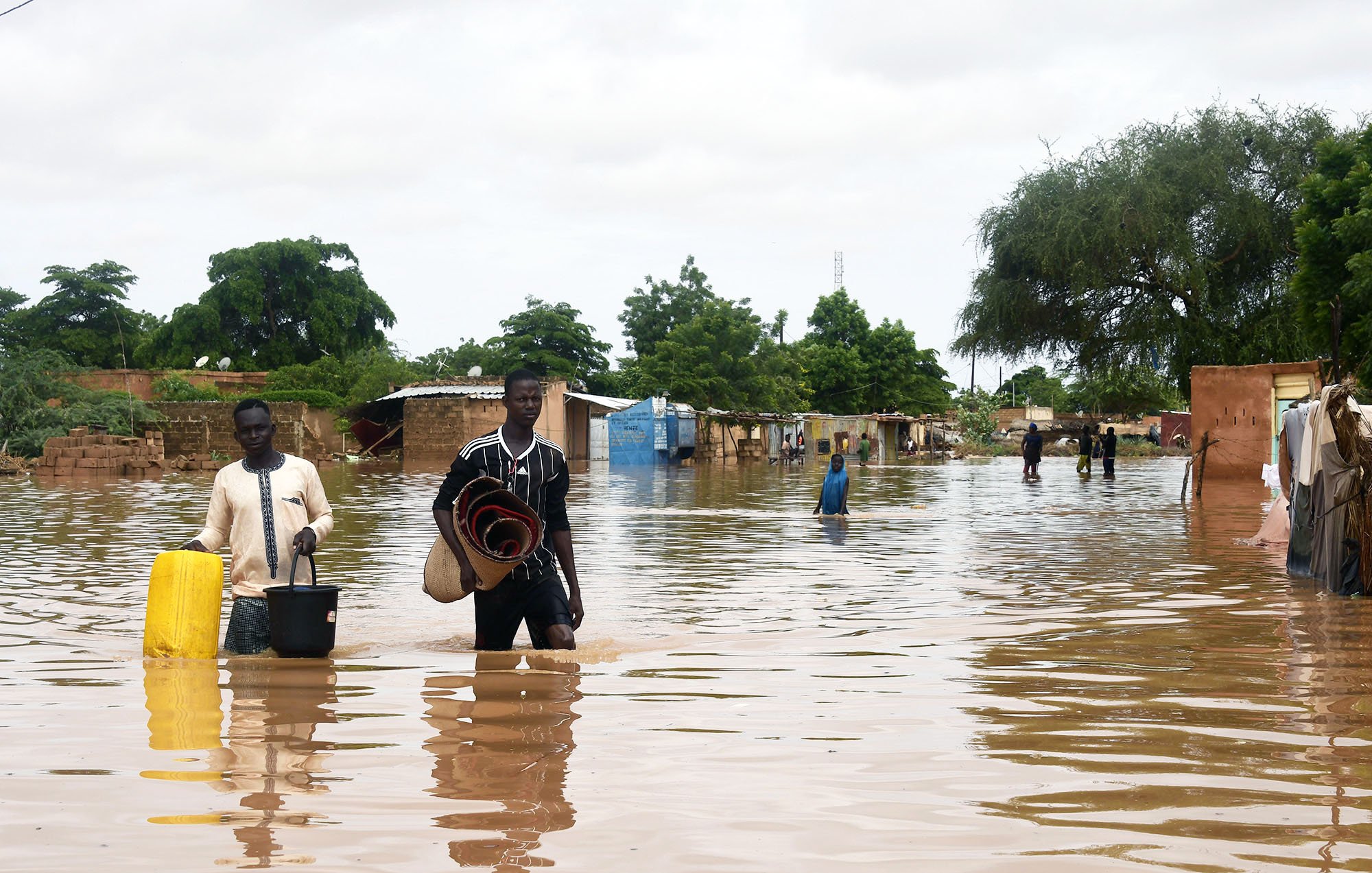 The image depicts a flooded area, with individuals wading through water that has inundated the surroundings. The water reaches knee-deep, affecting homes and structures in the background, which appear to be partially submerged. People in the scene are carrying various items, suggesting they're either evacuating or salvaging belongings. Trees and greenery can be seen around the area, indicating that it is a rural or semi-urban environment. The sky is overcast, contributing to the somber atmosphere of the flooding situation.