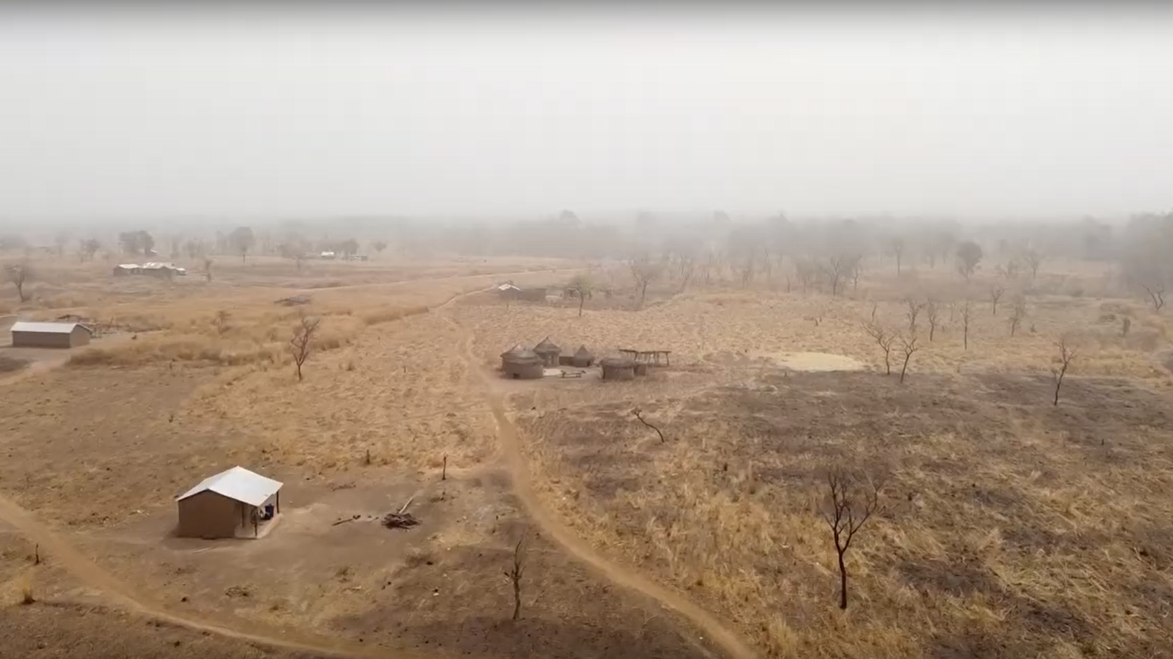 The image depicts a rural landscape characterized by dry, barren land. There are patches of brown grass and sparse vegetation, indicating an arid environment. In the foreground, a small structure with a white roof sits alongside a dirt path. In the background, a few more buildings can be seen, scattered amidst the landscape. The atmosphere appears hazy, possibly due to dust or smoke, giving the scene a muted and subdued appearance. Sparse trees dot the area, and the overall setting suggests a remote, pastoral region.