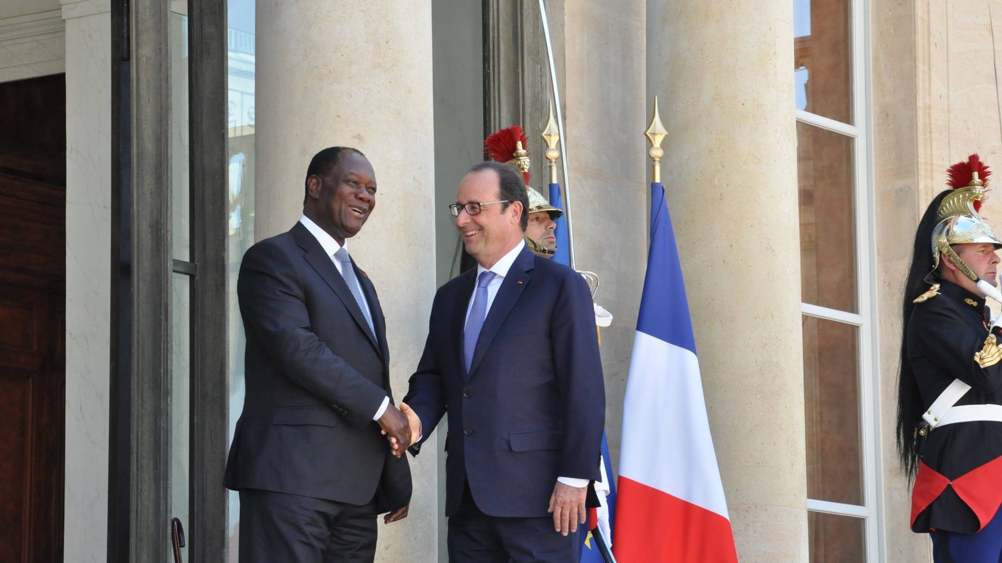The image shows two men shaking hands outside a large building, likely a government or official residence. One man is wearing a dark suit and the other is dressed in a suit and tie, likely representing different nations. Behind them, there is a large flag of France, and guards in traditional uniforms stand watch. The setting suggests a formal meeting or diplomatic engagement.