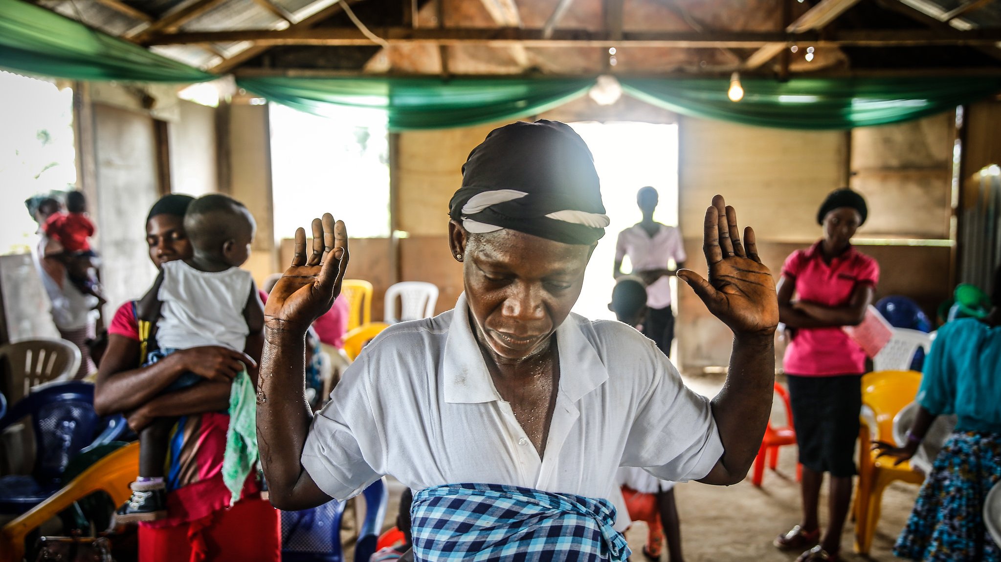 The image depicts a communal setting, likely a gathering or an event. In the foreground, a woman, dressed in a white top and a patterned shawl, raises her hands as if in prayer or meditation, demonstrating a moment of reverence or reflection. She appears focused and engaged in her spiritual practice. In the background, a variety of people are present, including individuals standing and others seated, indicating a diverse group participating in this activity. The interior of the space has wooden beams and a rustic charm, illuminated by soft lighting. The overall atmosphere seems to be one of community and spiritual connection.
