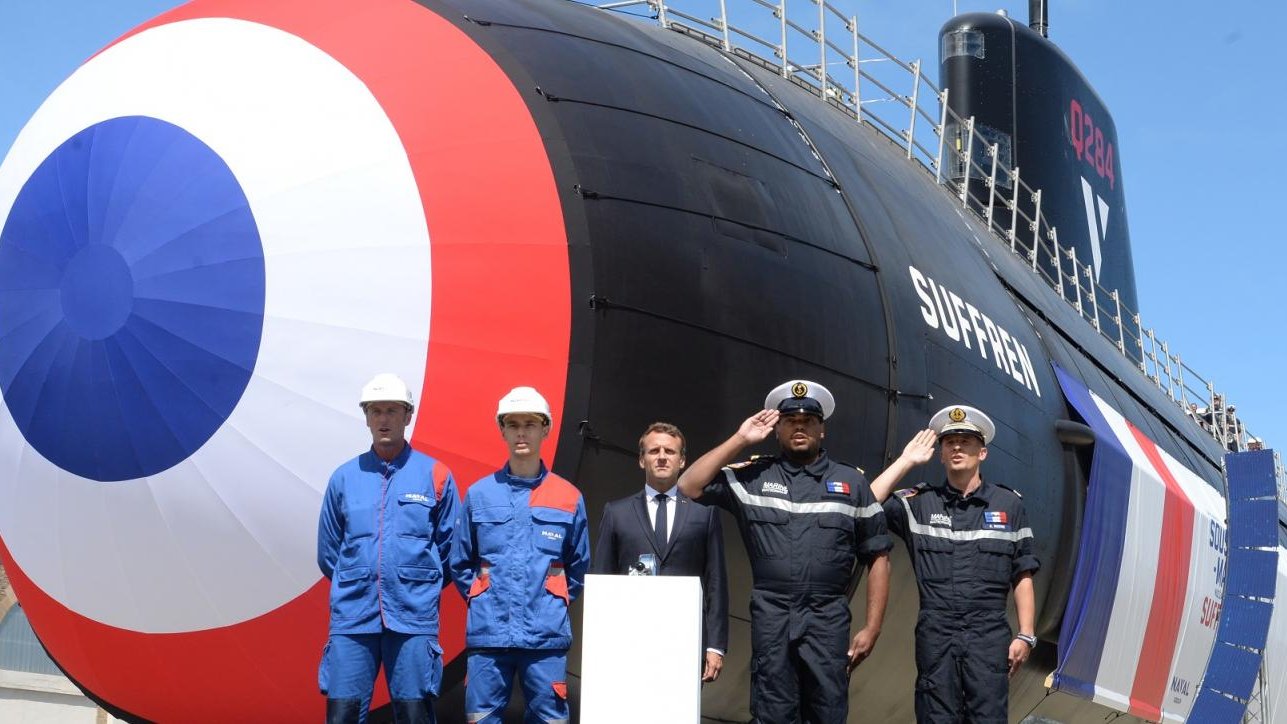 L'image représente un sous-marin de type Suffren, mis en avant lors d'une cérémonie en extérieur. Le sous-marin, aux couleurs caractéristiques bleu, blanc et rouge, se dresse majestueusement avec une grande coque stylisée. Le chiffre « 12 » est visible sur le côté. Devant le sous-marin, on voit plusieurs personnes en tenue de travail. À gauche, deux hommes portent des combinaisons bleues, un troisième homme, en costume, semble féliciter le projet, tandis que le quatrième, en uniforme, adopte une posture militaire. L'arrière-plan est dégagé avec un ciel bleu, ce qui donne une impression de journée ensoleillée. La scène évoque un moment de fierté et de célébration pour les employés et les responsables militaires, symbolisant un jalon important dans le domaine naval.