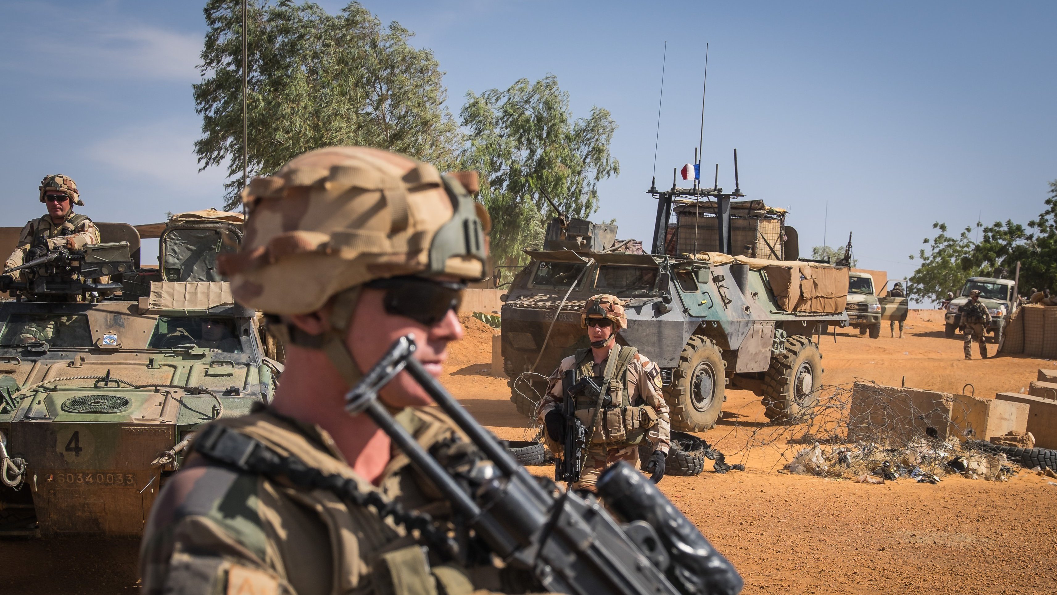 The image depicts a military scene in a desert environment. In the foreground, a soldier wearing a helmet and sunglasses is holding an assault rifle, looking serious and vigilant. Behind him, there are other soldiers and military vehicles, including an armored personnel carrier. The background shows a clear blue sky with some greenery and sand, indicating a remote and arid location. The overall atmosphere suggests a sense of readiness and military operations.