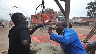 The image depicts a scene where two men are engaged in an activity involving a large sack. One man, wearing a blue coat, appears to be operating a scale or a weighing device to measure the weight of the sack being lifted by another man in a black shirt. The background shows a street with several structures and vehicles, suggesting a marketplace or an industrial area. There are also other people and containers visible in the background, contributing to the bustling atmosphere of the scene.