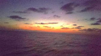 The image shows a serene seascape during twilight. The sky is transitioning from daylight to night, displaying a gradient of colors ranging from deep blue to orange near the horizon. Clouds are scattered across the sky, adding to the atmospheric depth. A single bright star is visible in the upper right portion of the image, contributing to the tranquil mood of the scene. The calm ocean reflects these colors, creating a peaceful and beautiful setting.