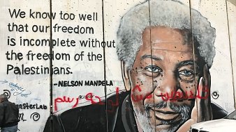 The image depicts a mural on a concrete wall, featuring a portrait of a man with gray hair and a thoughtful expression. The mural also includes a quote attributed to Nelson Mandela that reads, "We know too well that our freedom is incomplete without the freedom of the Palestinians." The background is a textured concrete surface, and there are some graffiti elements in red. The scene suggests themes of solidarity and freedom, particularly regarding the Palestinian struggle.