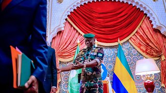The image shows a scene set in an ornate room. A military officer, dressed in camouflage with a green beret, is smiling and appears to be interacting with another individual. The background features decorative elements, including curtains and flags, likely representing Gabon or an official entity. There are also books and documents in the hands of a person standing to the left, indicating a formal context, possibly a meeting or ceremony. The overall atmosphere seems official and ceremonial.