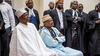 The image shows a group of individuals in formal attire, likely in a courtroom or legal setting. Two prominent figures in the foreground are seated, dressed in traditional attire. The person on the left wears a white outfit and a hat, while the individual on the right is in an aqua-colored garment with a distinctive headpiece. Behind them, several people are standing, some in legal robes, suggesting a gathering of lawyers or officials. The atmosphere appears serious and formal, indicating an important event or proceedings.