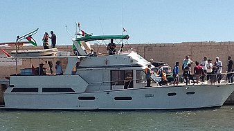 The image shows a large motorboat docked at a marina. The boat has a white hull with a green and white canopy on top. A group of people can be seen on the boat and along the dock, some engaging in conversation and others preparing to board. The surrounding area appears sunny, and there are flags displayed on the boat and nearby structures. The water is calm, reflecting the bright blue sky.