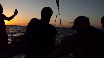 The image shows a group of people on a boat at sunset. The sky is painted in warm hues of orange and yellow as the sun sets on the horizon. Two people are visible, sitting with their backs to the camera, seemingly enjoying the view. One person is standing and holding a phone, possibly taking a photo. The water appears calm, reflecting the colors of the sky. The overall atmosphere is serene and relaxed.