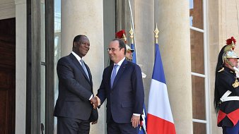 The image shows two men shaking hands outside a large building, likely a government or official residence. One man is wearing a dark suit and the other is dressed in a suit and tie, likely representing different nations. Behind them, there is a large flag of France, and guards in traditional uniforms stand watch. The setting suggests a formal meeting or diplomatic engagement.