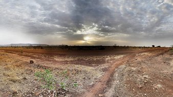 The image features a vast landscape under a cloudy sky, with the sun partially obscured by clouds, casting a soft light across the scene. The ground is dry and dusty, with patches of bare earth and sparse vegetation, including small plants and grasses. In the foreground, a path or trail diverges, leading into the distance, suggesting a sense of exploration in this open, natural environment. The overall tone is tranquil and somewhat desolate, evoking a sense of solitude in nature.