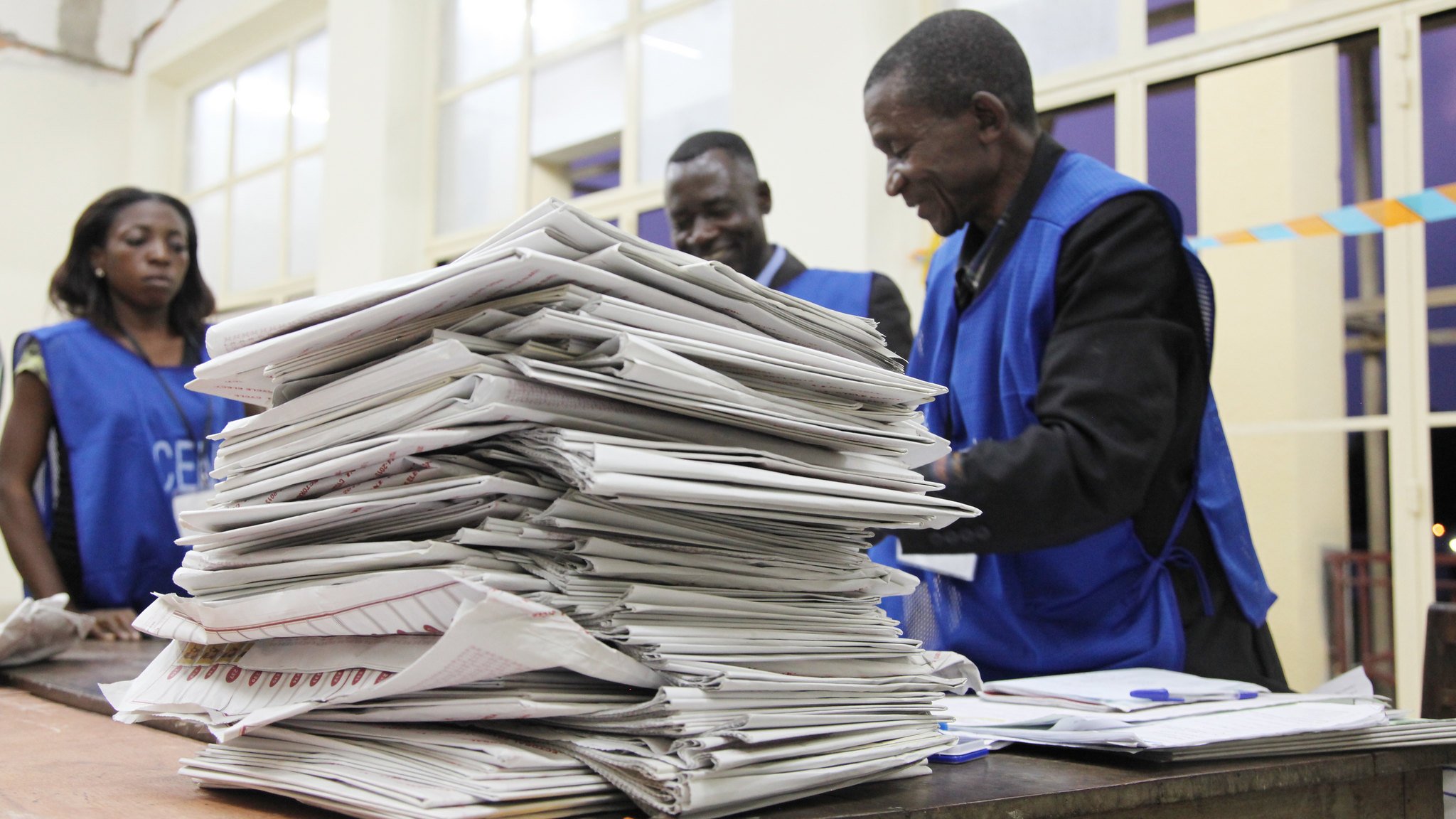 L'image montre une scène à l'intérieur d'un bureau de vote. Au premier plan, on aperçoit une grande pile de documents, probablement des bulletins de vote ou des formulaires électoraux, soigneusement empilés sur une table en bois. À l'arrière-plan, trois personnes en gilets bleus se trouvent près de la table, semblant discuter ou examiner les papiers. L'atmosphère est concentrée, suggérant une activité importante liée à un scrutin. La lumière éclaire la pièce, créant une ambiance sérieuse et professionnelle.