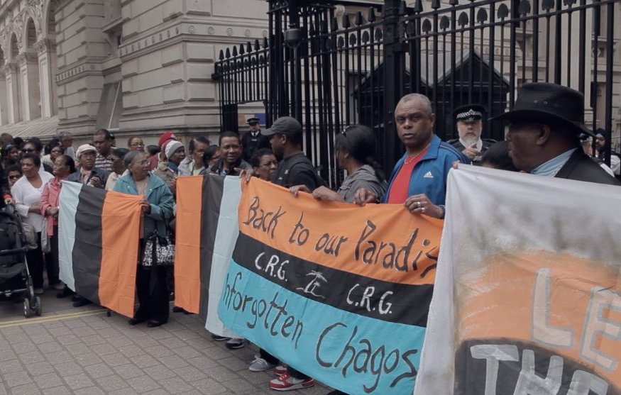 Dans cette image, un groupe de manifestants se rassemble devant un édifice imposant, probablement un bâtiment gouvernemental. Les personnes sont vêtues de vêtements variés, et certaines tiennent des pancartes. L'une des pancartes est colorée en orange, noir et bleu, et porte des inscriptions comme "Back to our Paradise" et "Forgotten Chagos". L'atmosphère est empreinte de détermination, avec des visages sérieux et engagés. Au fond, on peut voir des policiers surveillant la situation. L'environnement est urbain, avec des pavés au sol et des murs architecturaux ornés.