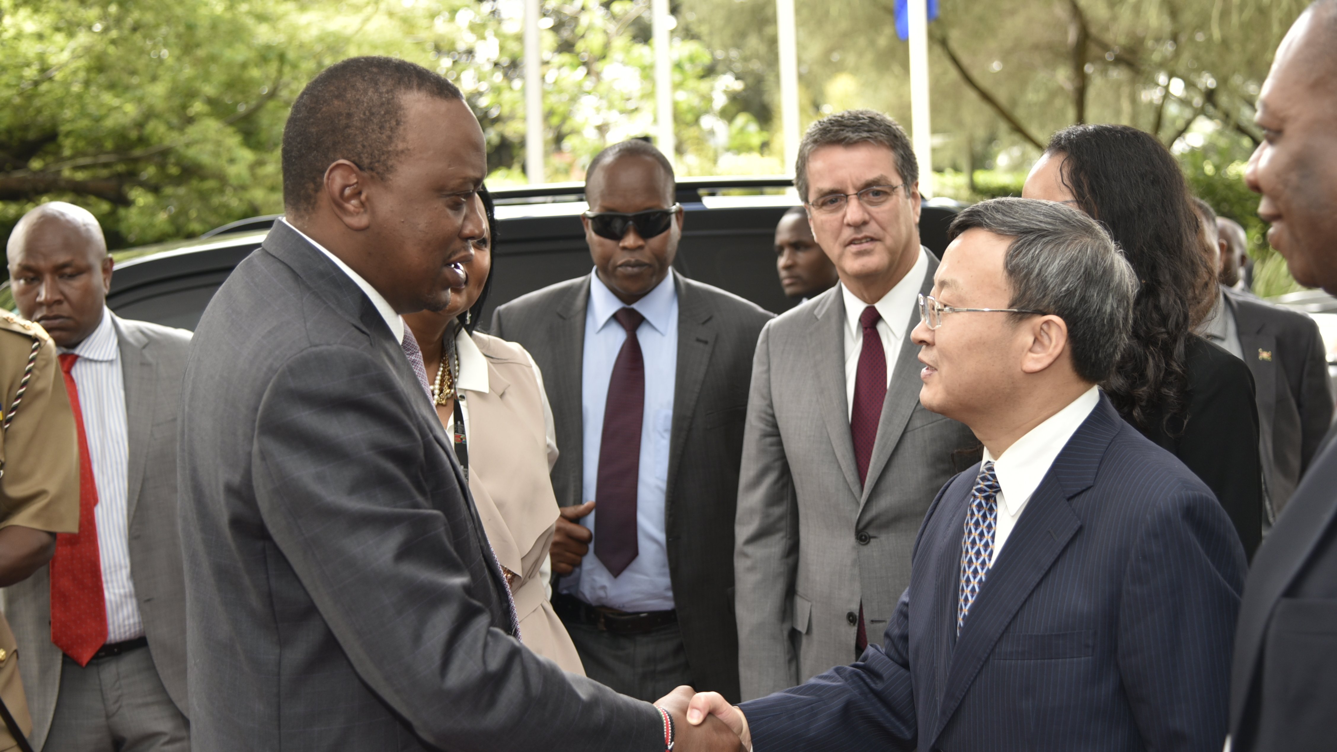 L'image montre une scène de rencontre officielle à l'extérieur, dans un environnement verdoyant avec des arbres et des drapeaux en arrière-plan. Au centre, deux hommes se serrent la main. L'un est habillé en costume sombre, avec une expression sérieuse, tandis que l'autre porte une cravate et un costume clair. En arrière-plan, d'autres personnes observent la rencontre, certaines avec des lunettes de soleil, indiquant un moment formel. L'atmosphère est celle d'une discussion diplomatique.