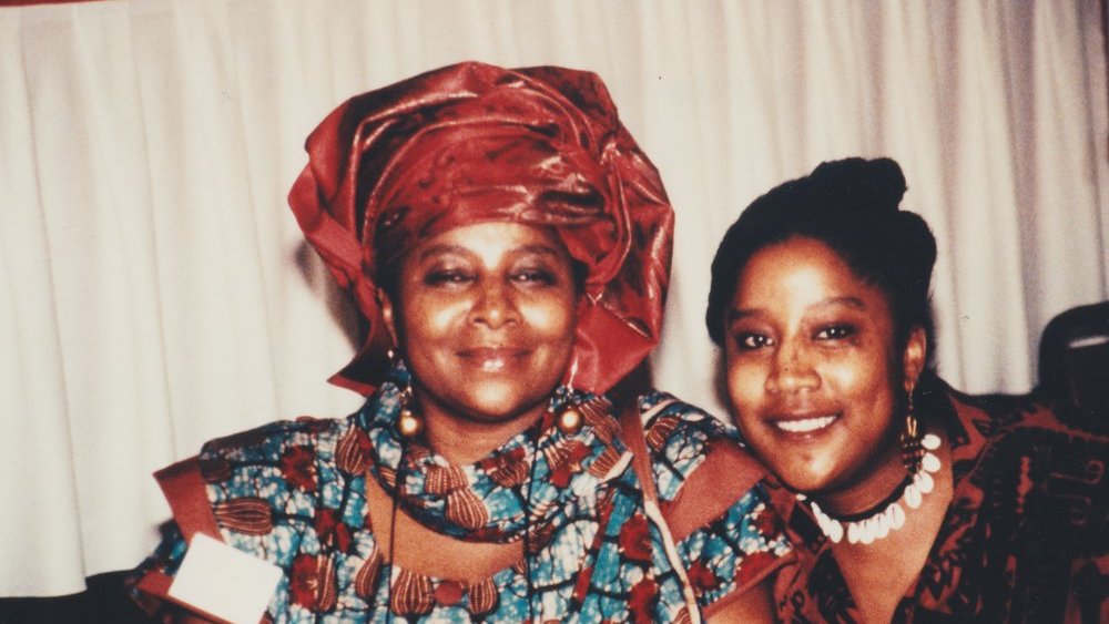 L'image montre deux femmes souriantes posant côte à côte. La femme à gauche porte un vêtement traditionnel aux motifs colorés, accompagné d'un foulard assorti. Sa expression est douce et chaleureuse. À sa droite, la seconde femme affiche également un grand sourire ; elle porte un haut à motifs et des accessoires qui ajoutent une touche élégante à son apparence. L'environnement semble être festif, avec une nappe blanche en arrière-plan, laissant planer une atmosphère accueillante et joyeuse.