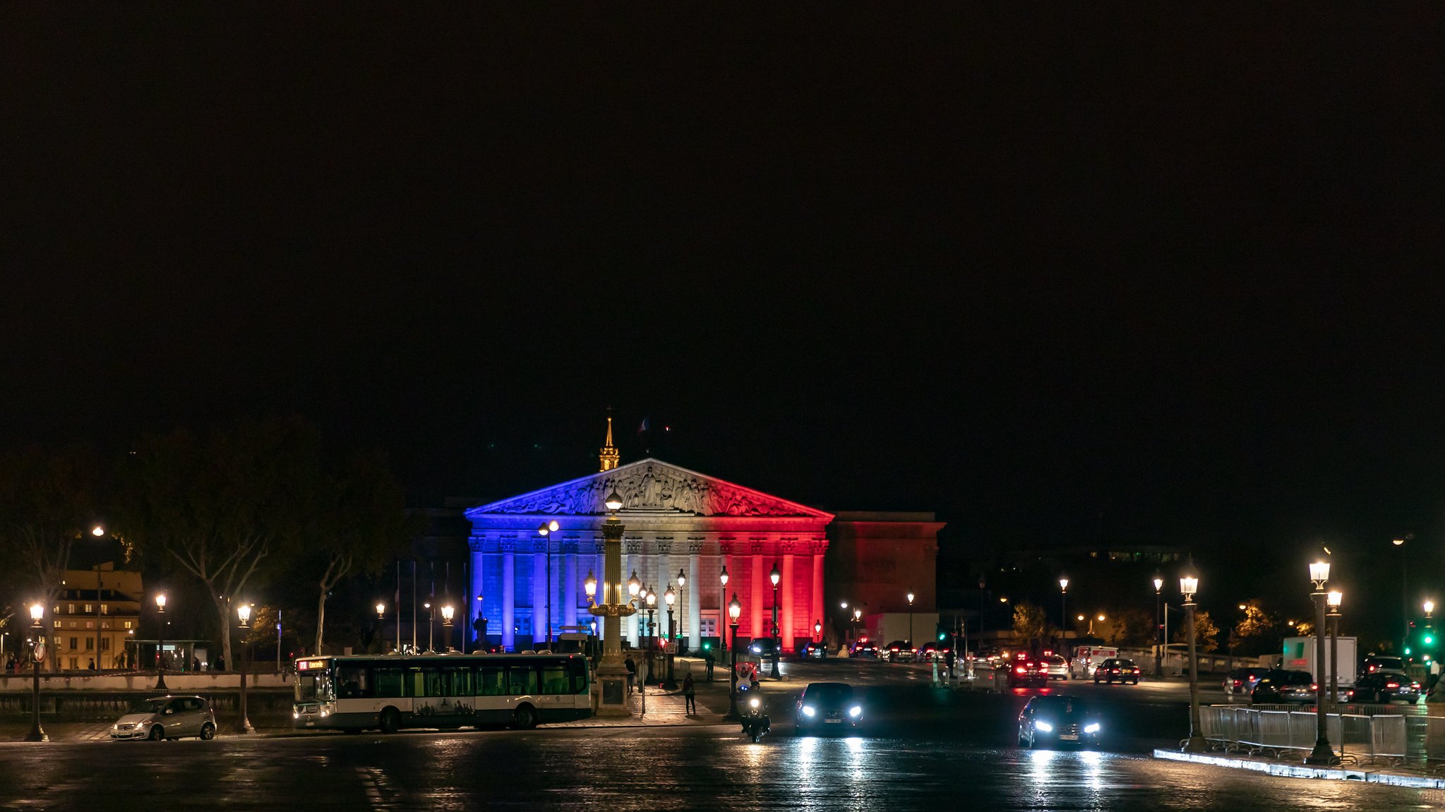 Imaginez un bâtiment majestueux illuminé par des lumières tricolores. La façade est peinte de bleu, de blanc et de rouge, évoquant les couleurs du drapeau français. Le ciel nocturne est sombre, créant un contraste saisissant avec les lumières vibrantes. À l'avant, vous pouvez sentir la présence d'une place animée, avec des voitures qui circulent et des lampadaires qui diffusent une lumière douce sur le pavé. La scène dégage une ambiance festive et patriotique, symbolisant la fierté nationale.