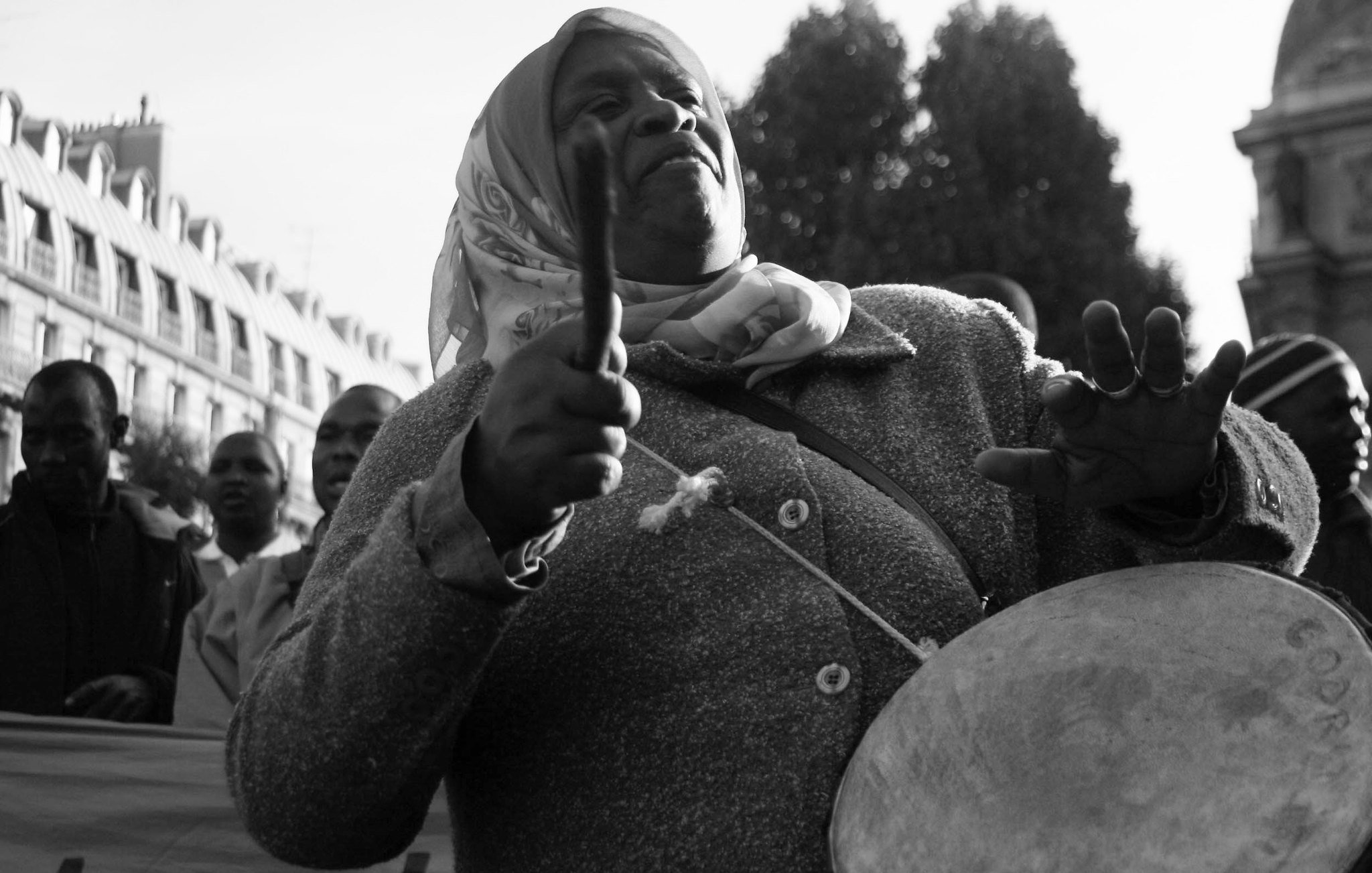 L'image représente une femme engagée dans un moment de mobilisation. Elle porte un manteau gris et un foulard autour de la tête. Ses bras sont levés, tenant des baguettes, et elle semble jouer d'un instrument de percussion, probablement un tambour. Son visage exprime une concentration et une passion profonde, alors qu'elle fait vibrer la musique au sein d'une foule. En arrière-plan, on peut distinguer d'autres personnes, suggérant une manifestation ou un rassemblement collectif. L'atmosphère est chargée d'énergie et de détermination. La photo est en noir et blanc, ce qui ajoute une dimension intemporelle à cette scène de citoyenneté active.