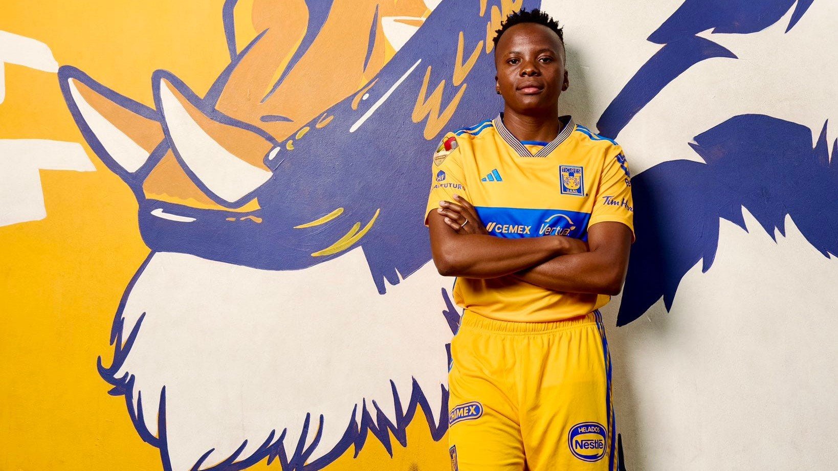 L'image montre un jeune homme debout devant un mur peint en jaune et bleu, qui représente un tigre rugissant. Il porte un uniforme de footbal aux couleurs vives, principalement jaune, avec des détails bleus. Son attitude est confiante ; il se tient les bras croisés, affichant une expression de détermination. Le mur qui l'entoure ajoute une dynamique visuelle avec des motifs de griffes et des éléments stylisés qui renforcent le thème du tigre. Le sol est recouvert d'herbe artificielle, évoquant un terrain de sport.