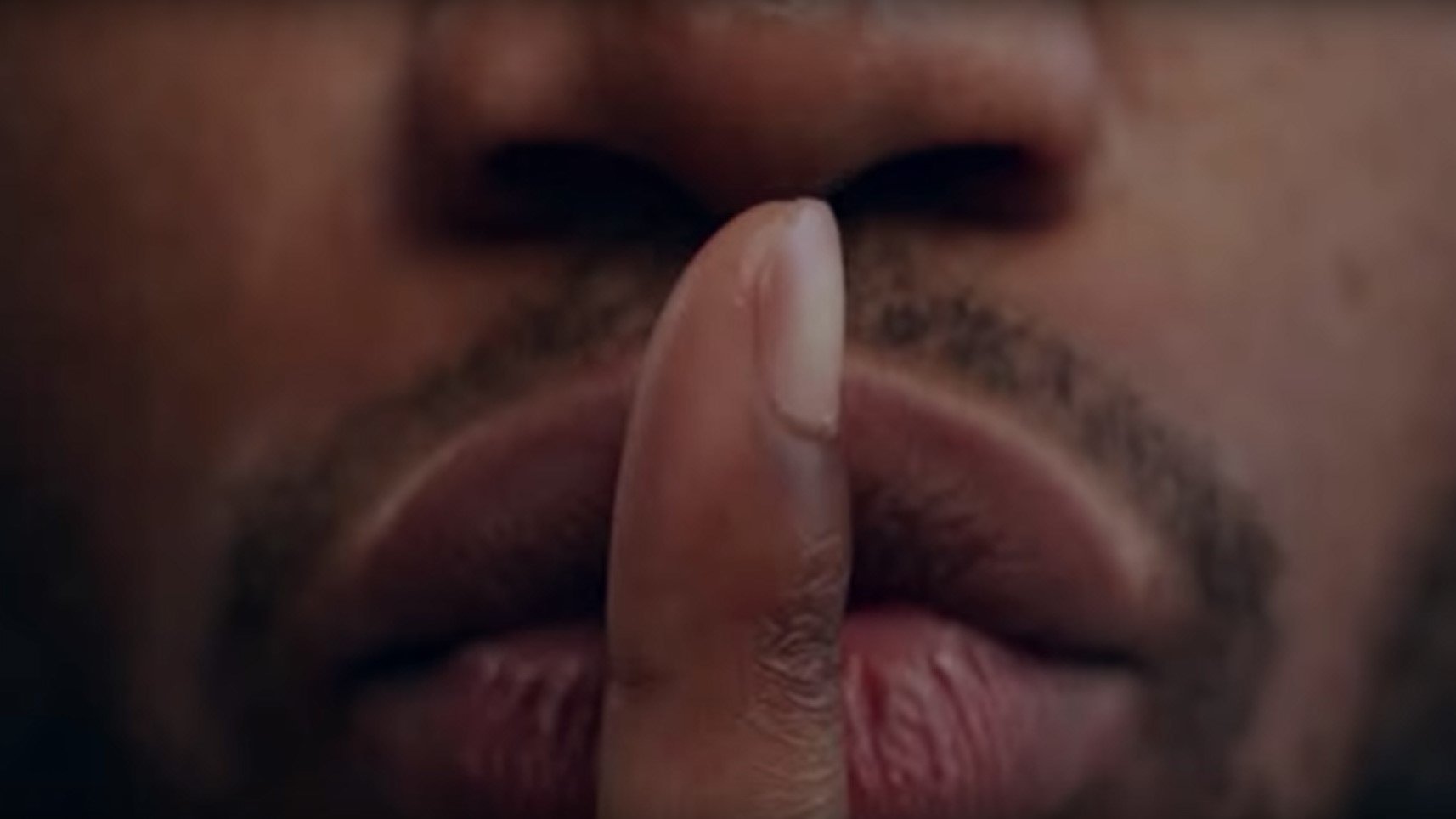 L'image présente un gros plan sur un visage, principalement la zone des lèvres et du nez. Un doigt est placé devant la bouche, formant le geste universel du « chut ». La peau est de teinte foncée, et les lèvres sont bien définies, affichant une légère brillance. Le regard de la personne est probablement dirigé vers l'extérieur de l'image, transmettant une impression de mystère ou de secret. L'arrière-plan est flou, ce qui met l'accent sur le geste et l'expression du visage.
