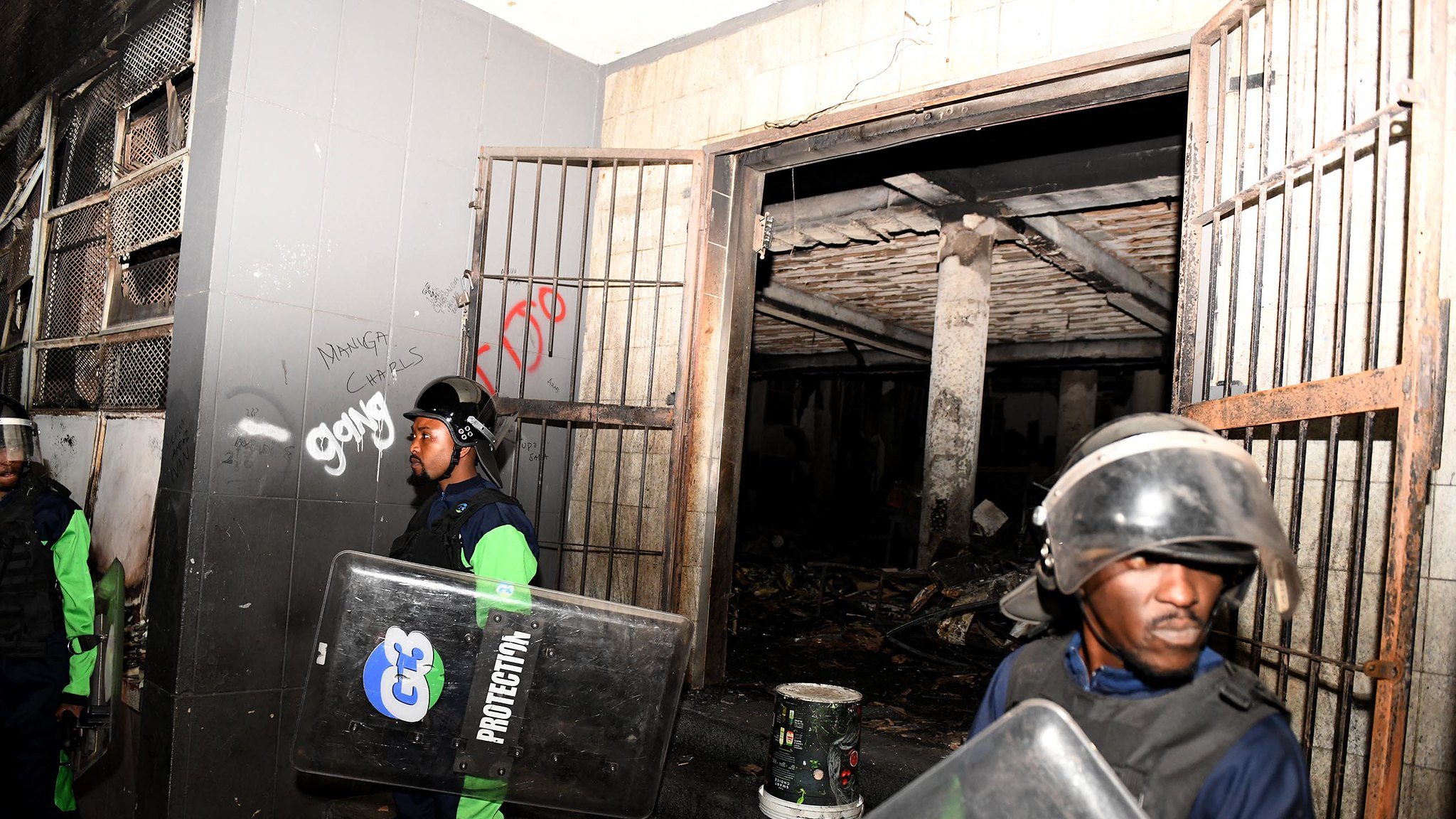 L'image montre une scène de sécurité, probablement après un événement perturbateur. On y voit deux agents de sécurité, vêtus d'un uniforme bleu et vert, portant des casques de protection et tenant des boucliers. Ils se tiennent devant une entrée, à côté de portes métalliques ouvertes. À l'intérieur, on aperçoit un espace sombre avec des traces de dégâts, comme des murs noircis. L'environnement semble chaotique, avec des débris visibles au sol. Des graffitis sont présents sur les murs, ajoutant une touche de désordre à la scène. L'atmosphère générale dégage un sentiment de tension et de vigilance.