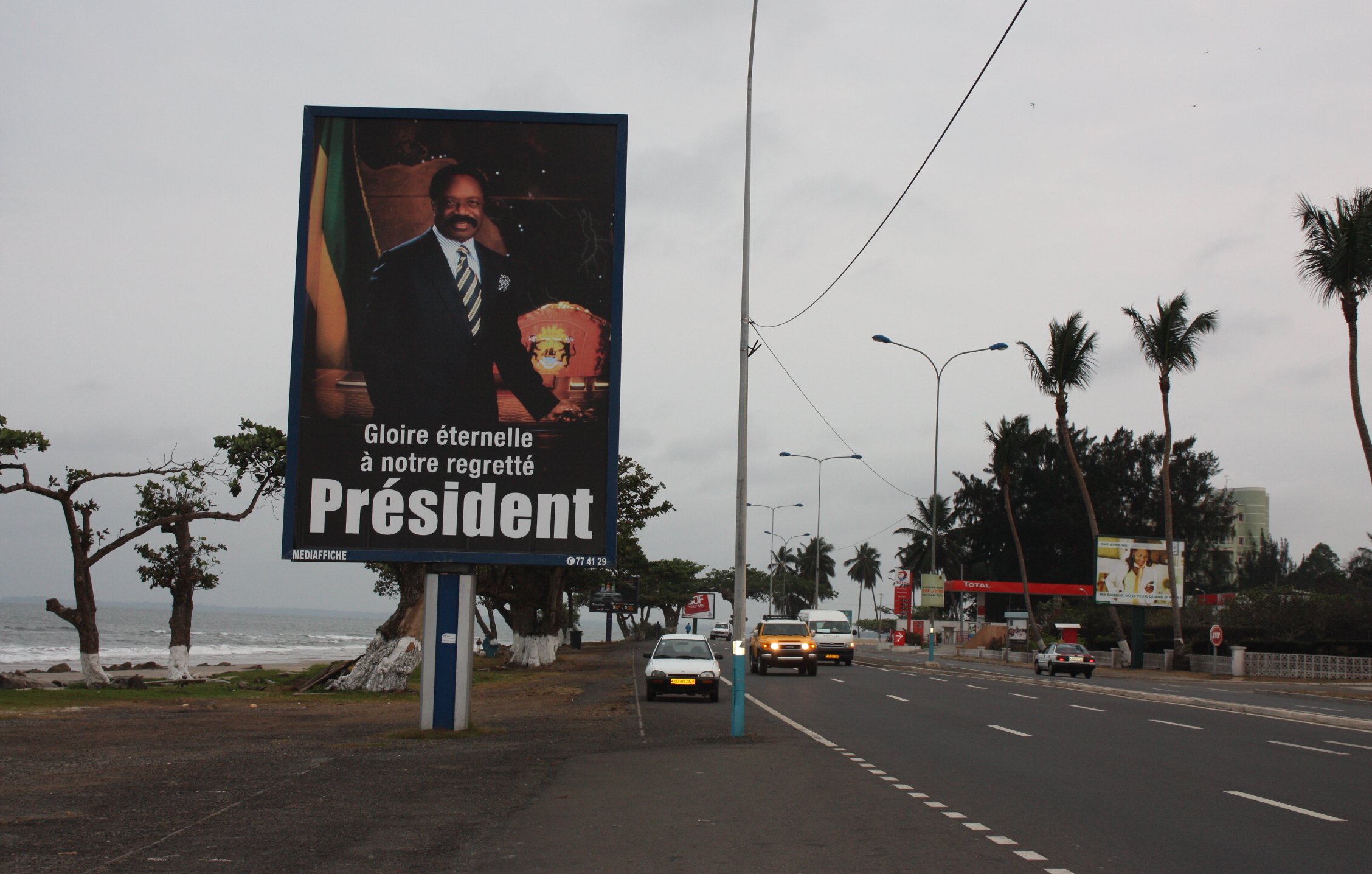 L'image montre un grand panneau publicitaire sur le bord d'une route. Ce panneau présente le portrait d'un homme en costume, avec un air solennel, tenant un insigne ou un symbole de pouvoir. En dessous de son image, il y a un texte qui dit : "Gloire éternelle à notre regretté Président", exprimant un hommage à un dirigeant décédé. En arrière-plan, on peut apercevoir une route avec quelques voitures et des palmiers, ainsi qu'une plage visible à proximité. Le ciel est nuageux, ce qui donne une ambiance mélancolique à la scène.
