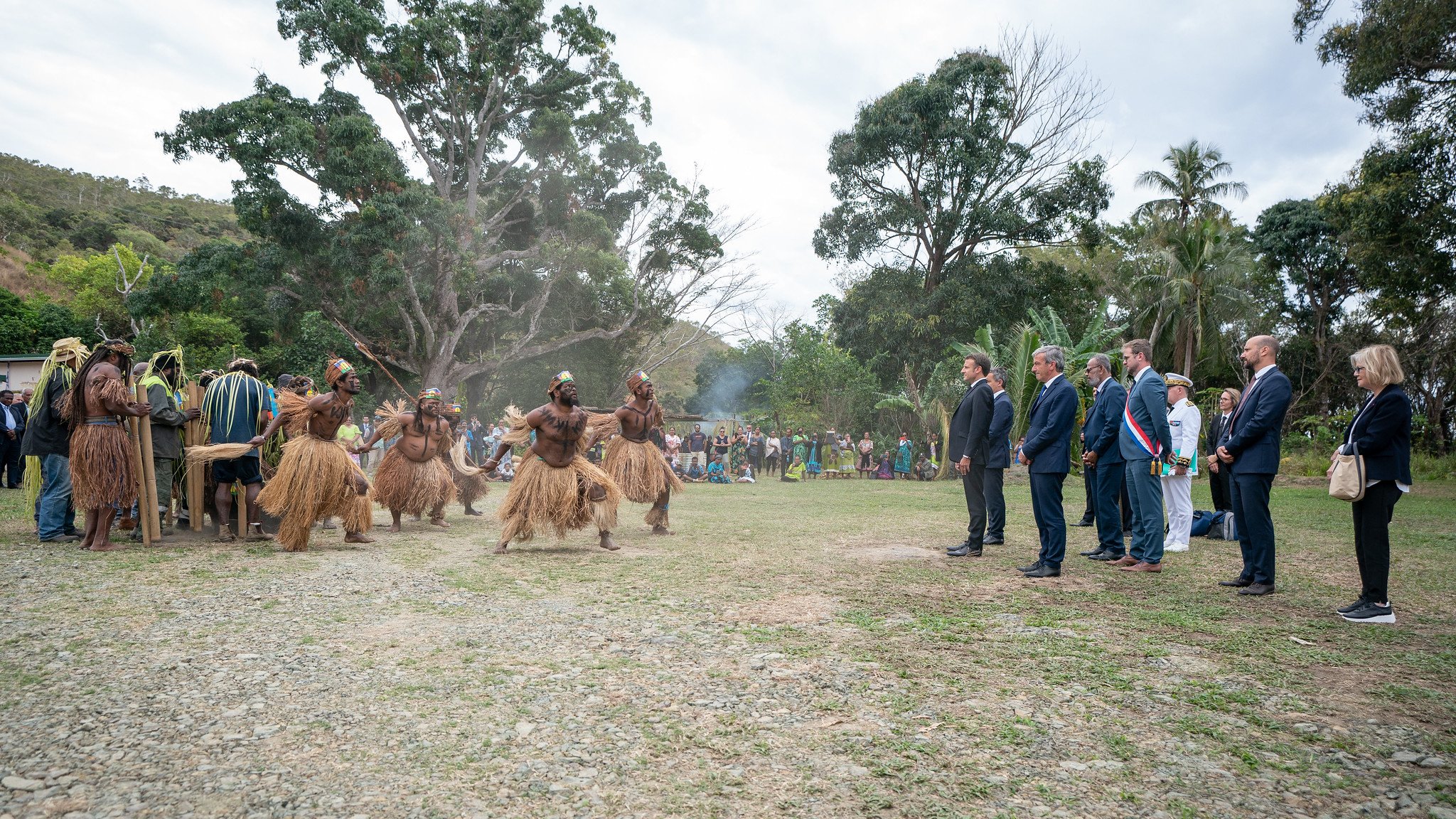 L'image montre un groupe de personnes en tenue traditionnelle, qui semblent participer à une cérémonie ou une célébration. Au centre, des hommes vêtus de costumes en fibres naturelles dansent avec énergie. Ils sont entourés par un public, composé de personnes en tenue formelle, qui observe attentivement la scène. L'environnement est verdoyant, avec des arbres et une légère brume qui donne une atmosphère presque mystique. À l'arrière-plan, un groupe de spectateurs semble apprécier l'événement, ajoutant une dimension communautaire à cette célébration culturelle.