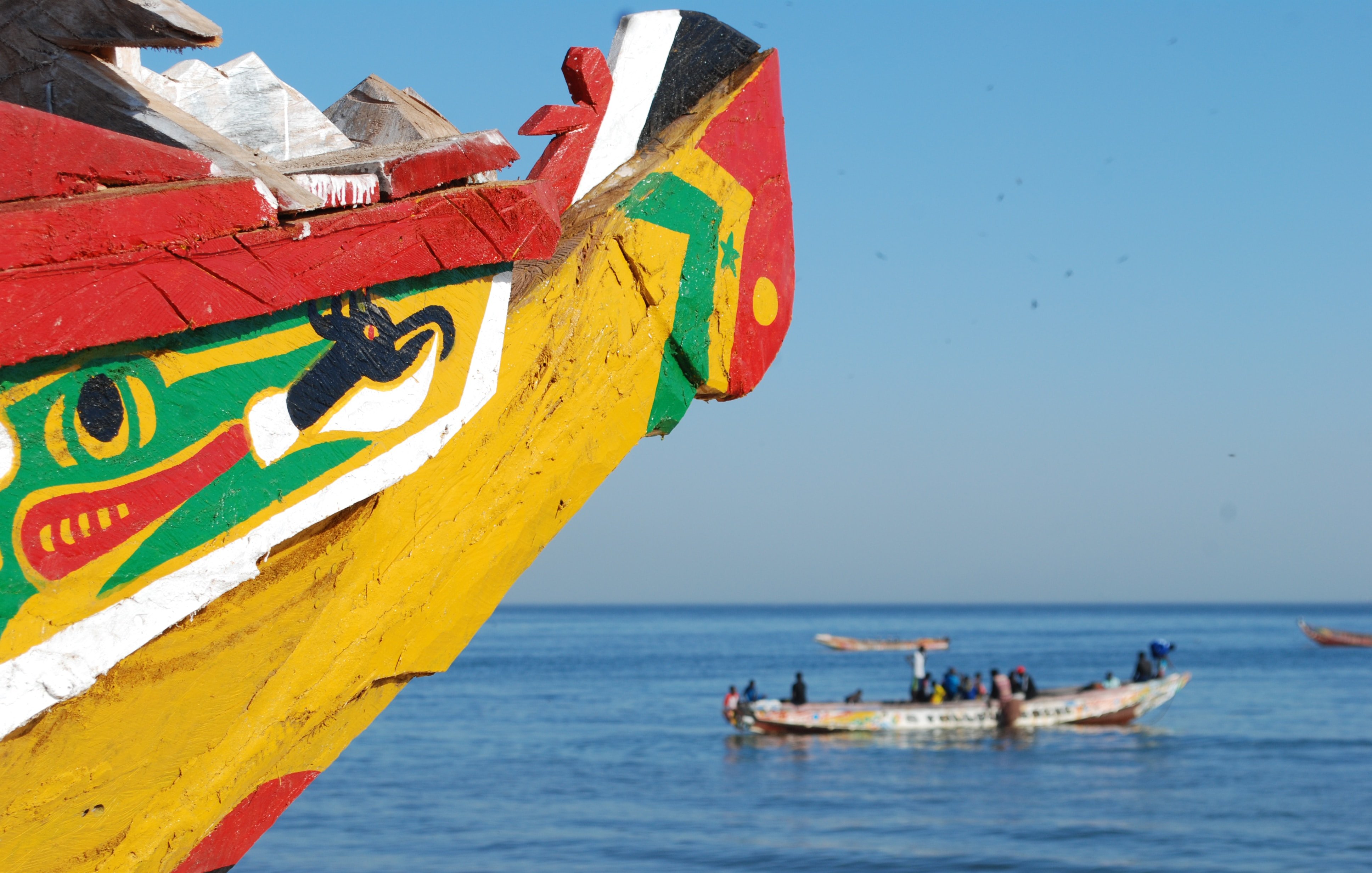 Cette image représente la proue colorée d'une pirogue traditionnelle, peinte de motifs vifs dans des couleurs comme le rouge, le jaune, et le vert. Au loin, on peut apercevoir d'autres bateaux de pêche qui naviguent paisiblement sur l'eau calme d'une mer bleue. Le ciel est dégagé, créant une atmosphère sereine et ensoleillée. L'air salin doit être frais, et on peut entendre le bruit des vagues et peut-être des conversations des marins qui travaillent sur leurs embarcations. L'ensemble dégage une ambiance maritime typique des côtes africaines.
