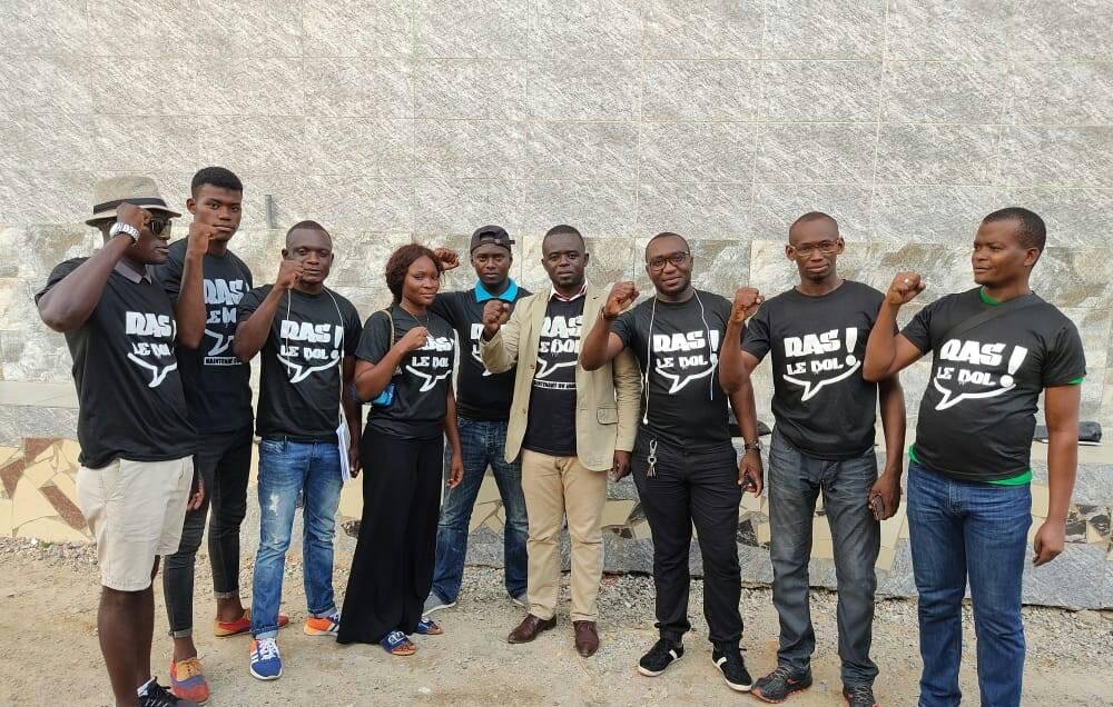 L'image montre un groupe de onze personnes rassemblées, certaines portant des t-shirts noirs avec des inscriptions. Ils affichent tous des expressions déterminées et se tiennent debout, avec quelques-uns levant le poing en signe de solidarité ou de protestation. Les membres du groupe sont de différents sexes et portent des vêtements variés, allant de t-shirts décontractés à des vêtements plus formels. L'arrière-plan est constitué d'un mur en pierre, ajoutant une texture neutre à la scène. L'ambiance semble énergique et engagée.