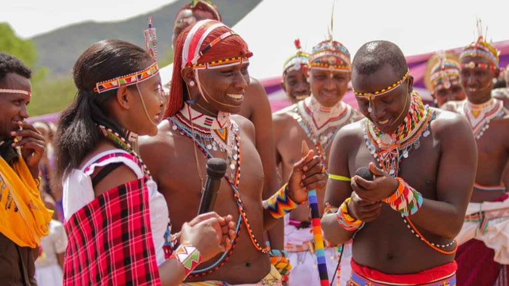 Dans cette image, un groupe de personnes participe à une cérémonie culturelle vibrante. Au premier plan, une femme, vêtue d'une robe traditionnelle colorée avec des motifs, tient un micro et s'adresse à un homme qui se tient à côté d'elle. Cet homme porte un pagne traditionnel et est orné de nombreux accessoires en perles. Ils sont tous deux entourés de plusieurs autres hommes, également en tenue traditionnelle, qui sourient et semblent s'engager dans une conversation joyeuse. Les vêtements sont décorés de motifs colorés et de bijoux en perles, ce qui évoque une atmosphère festive. L'arrière-plan montre des collines verdoyantes, et l'ambiance générale dégage un sens de communauté et de célébration de la culture. L'image respire l'énergie et la convivialité, illustrant des rituels ou des danses traditionnelles.