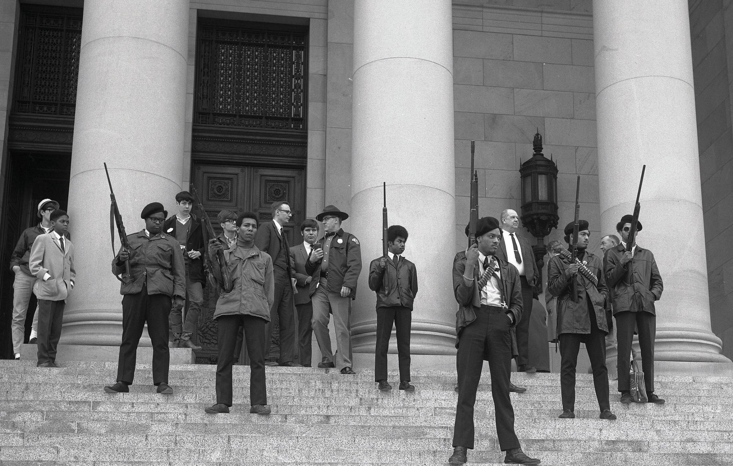 L'image montre un groupe de personnes sur les marches d'un bâtiment imposant, probablement une institution gouvernementale. Au premier plan, plusieurs jeunes hommes se tiennent debout, portant des vestes sombres et des armes à feu, avec des expressions déterminées sur leurs visages. Ils sont entourés d'autres individus, certains vêtus de manière formelle, comme des costumes et des chapeaux. Tous semblent engagés dans une sorte de manifestation ou de rassemblement, émettant un sentiment de tension et de détermination. L'architecture du bâtiment est majestueuse, avec de grandes colonnes en arrière-plan. L'atmosphère générale de l'image évoque une période de lutte pour les droits civiques.