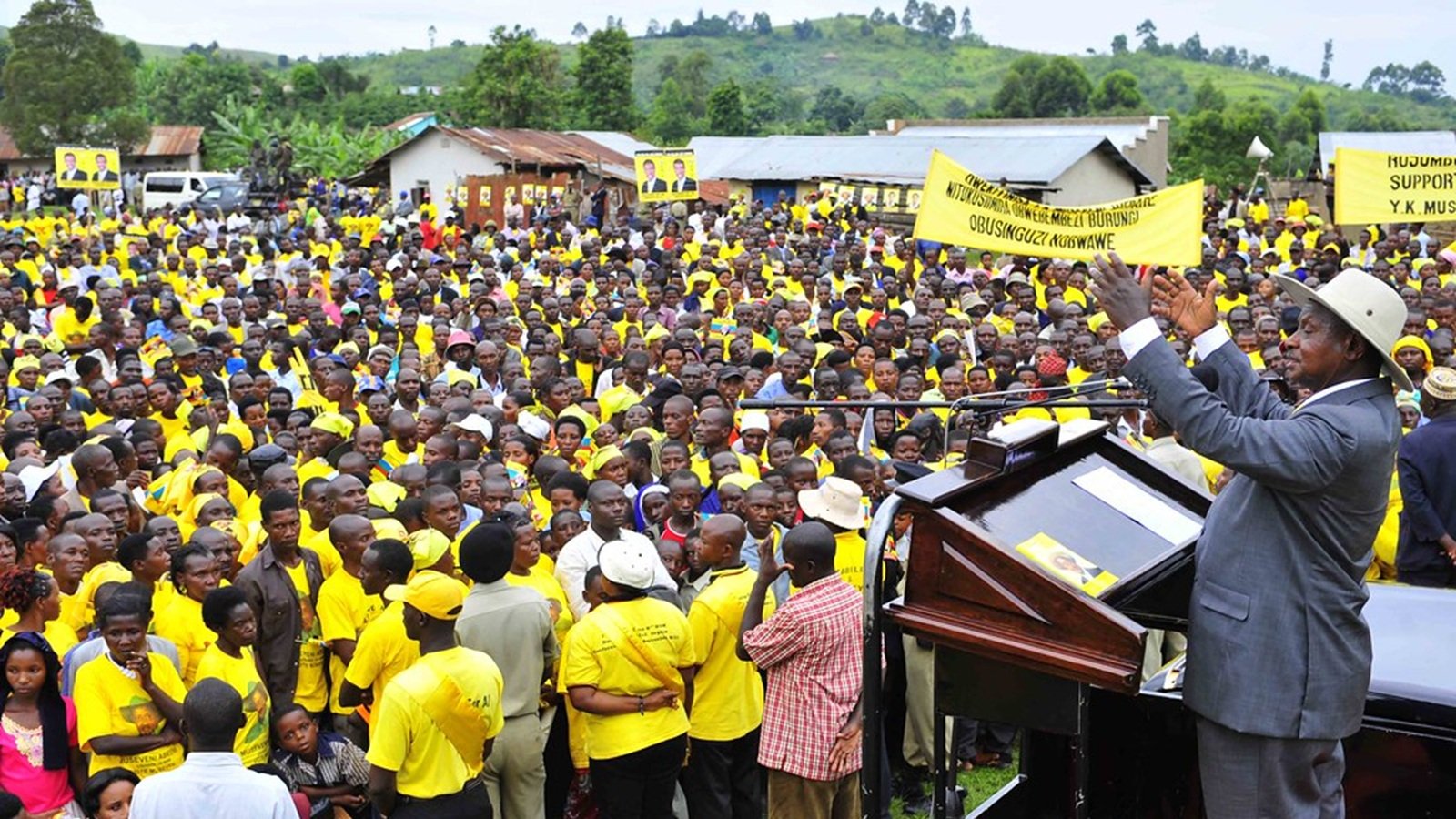 L'image montre une grande foule rassemblée en plein air. Les personnes dans la foule portent majoritairement des vêtements jaunes, ce qui pourrait indiquer qu'elles soutiennent un mouvement ou un parti spécifique. Au premier plan, un homme, vraisemblablement un orateur ou un dirigeant, se tient derrière un podium. Il lève les bras, semblant interagir avec la foule et exprimer un message puissant. En arrière-plan, on aperçoit des bâtiments simples et des collines verdoyantes, donnant un cadre naturel à l'événement. L'atmosphère semble énergique et mobilisée.