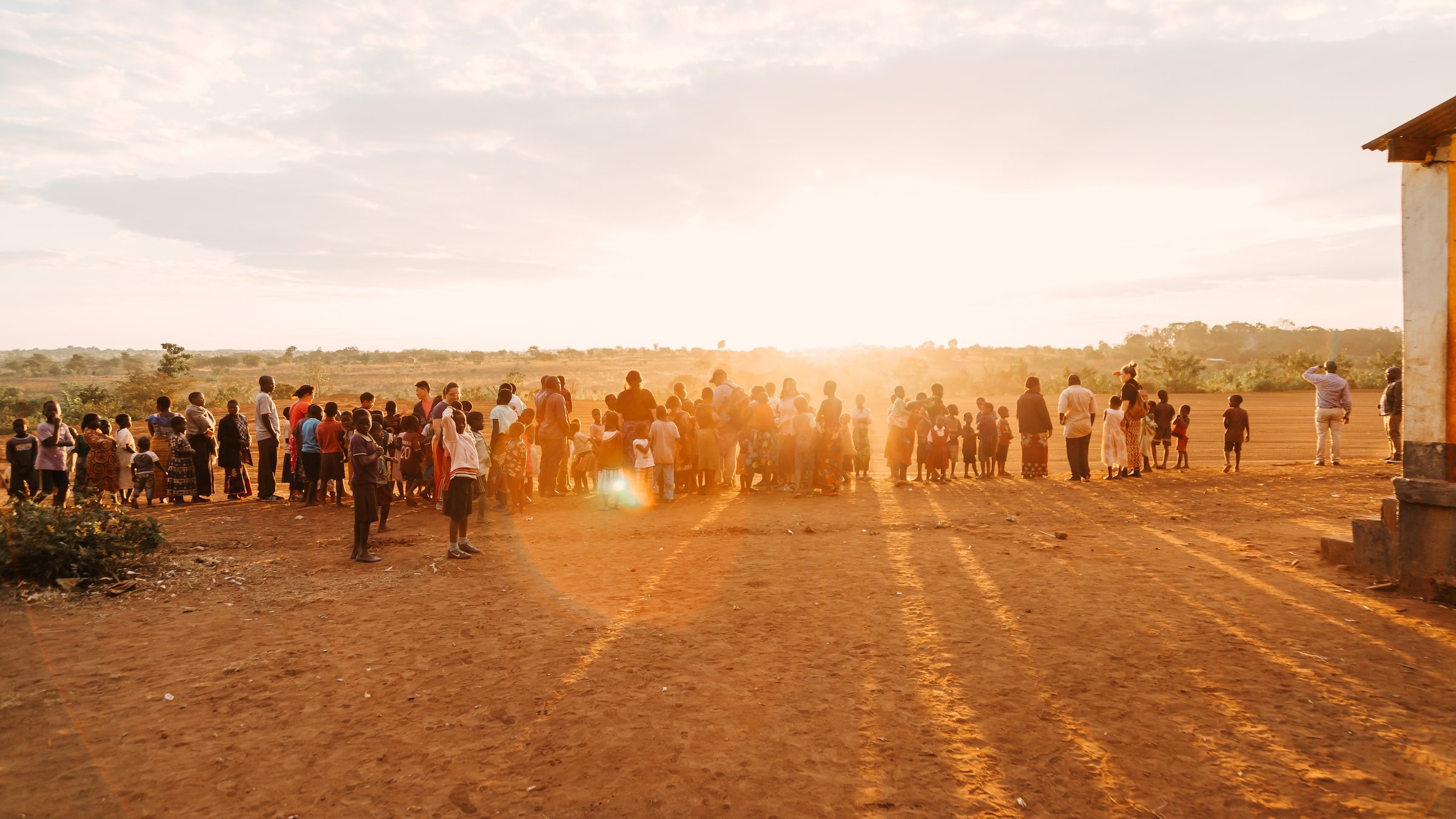 L'image présente une scène de rassemblement en plein air, au coucher du soleil. Au premier plan, un groupe nombreux de personnes se tient sur un terrain poussiéreux, probablement un village ou une communauté rurale. Les silhouettes des adultes et des enfants se distinguent distinctement alors qu'ils font face à l'horizon où le soleil descend, projetant de longues ombres sur le sol. L'arrière-plan est composé de plantes et d'une vaste étendue de paysage, créant une atmosphère paisible et chaleureuse. Le ciel, teinté de couleurs douces, suggère une fin de journée tranquille.