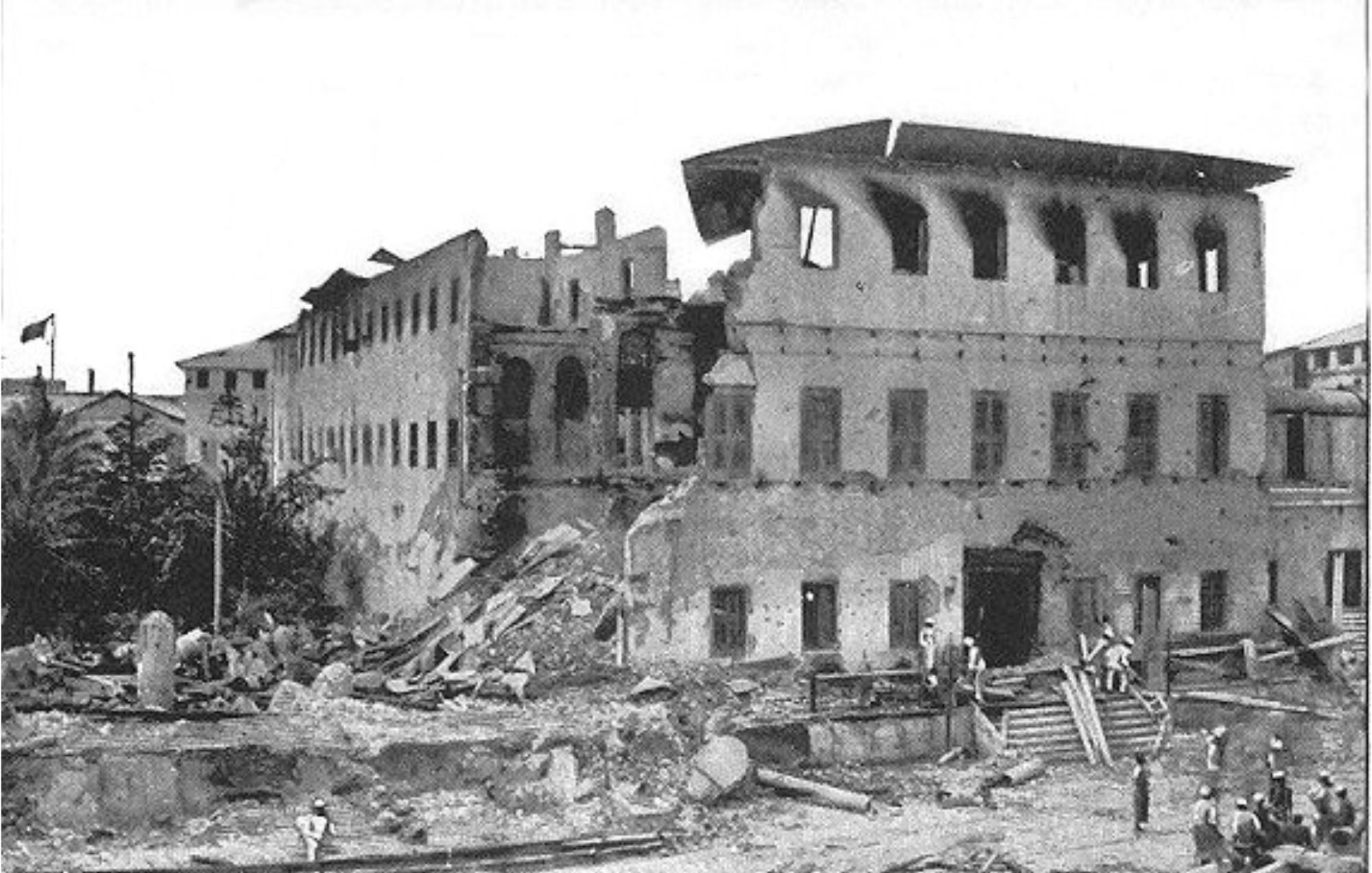 L'image montre un bâtiment sévèrement endommagé, probablement à la suite d'un tremblement de terre ou d'une catastrophe. La façade du bâtiment est partiellement effondrée, révélant des murs fissurés et des fenêtres brisées. Des débris sont éparpillés autour, créant un paysage chaotique. Au premier plan, on peut voir un groupe de personnes qui semblent examiner les dégâts, échangeant probablement leurs impressions. L'environnement est empreint d'une atmosphère de désolation, illustrant les conséquences d'un événement tragique sur l'architecture et la communauté environnante.