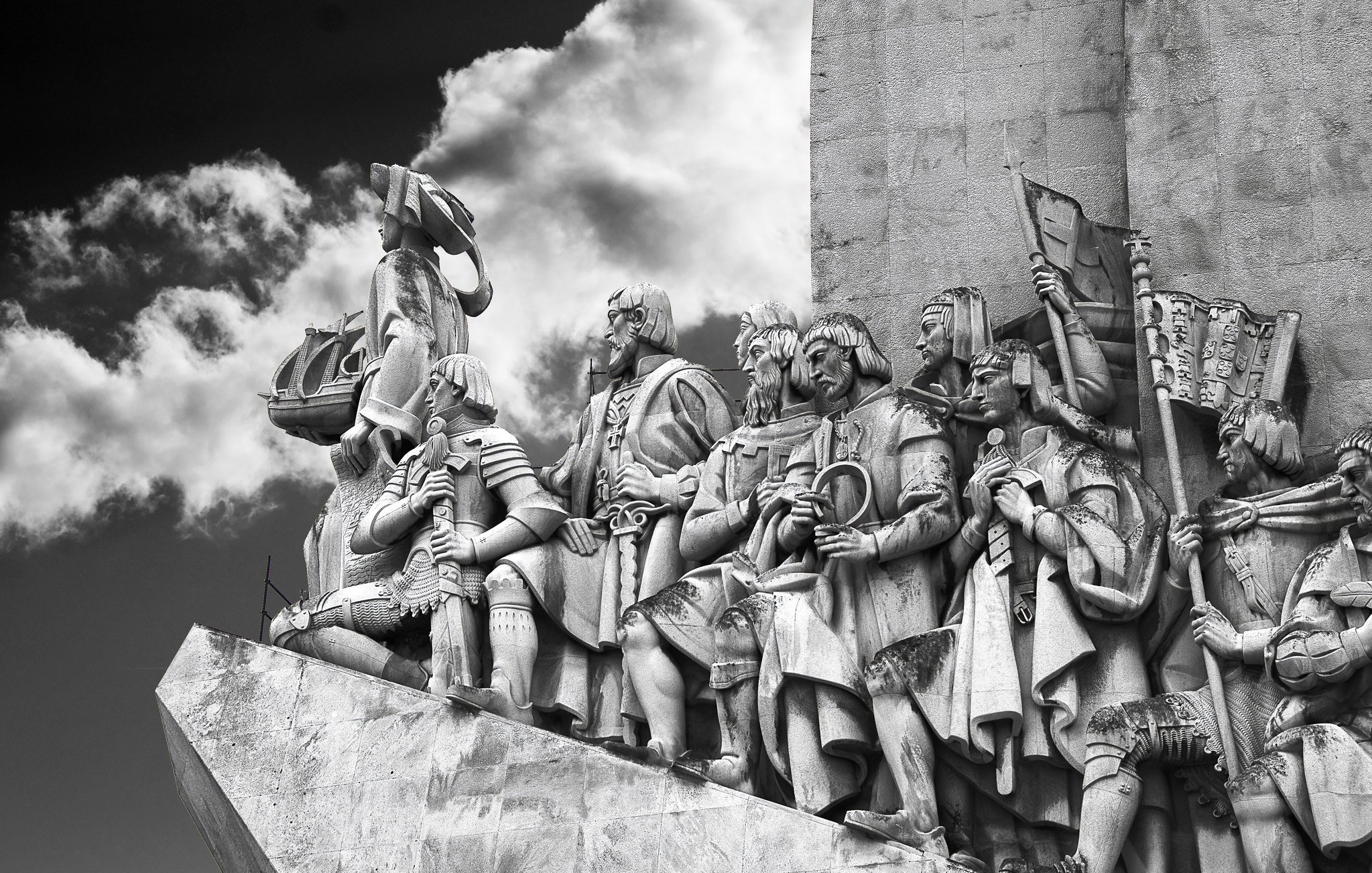 L'image représente un relief sculpté monumentale. On y voit une procession de figures humaines, probablement des explorateurs ou des conquérants, qui se tiennent debout sur une plateforme. Les personnages sont vêtus de costumes historiques et affichent des expressions déterminées. Certains d'entre eux portent des armes, des drapeaux et d'autres objets symboliques, témoignant de leurs exploits. En arrière-plan, on aperçoit des nuages sombres qui ajoutent une atmosphère dramatique à la scène. Le tout est sculpté dans un matériau clair, probablement de la pierre, permettant de bien contraster avec le ciel nuageux. Cette œuvre dégage une impression de puissance et de grandeur, évoquant une célébration de l'histoire et de l'aventure.